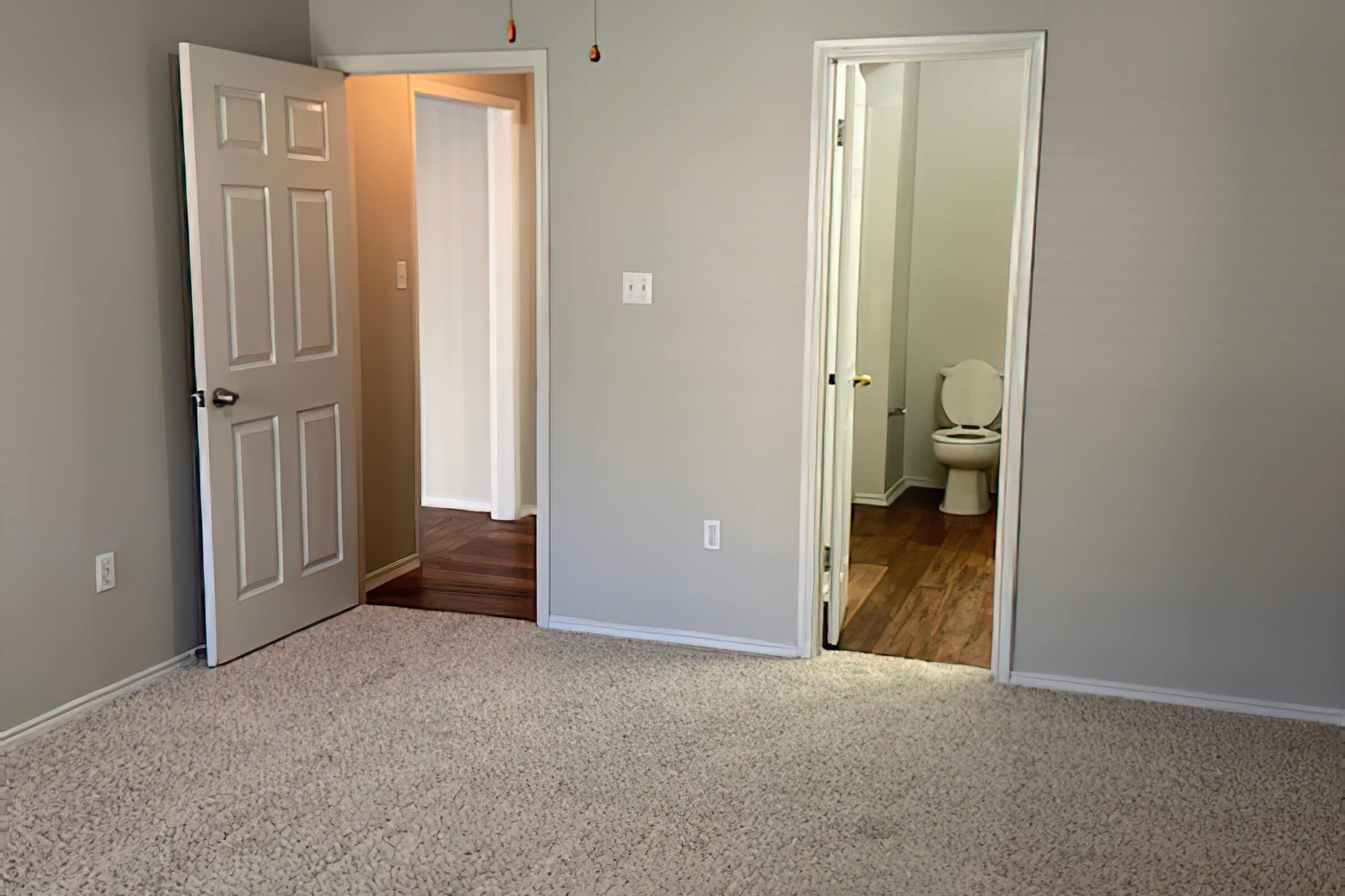 A light-colored room with beige carpet and gray walls. There are two doors: one leading to a bathroom with a toilet visible in the background, and another door slightly ajar leading to a hallway or another space. The lighting is bright and the room appears tidy and well-maintained.