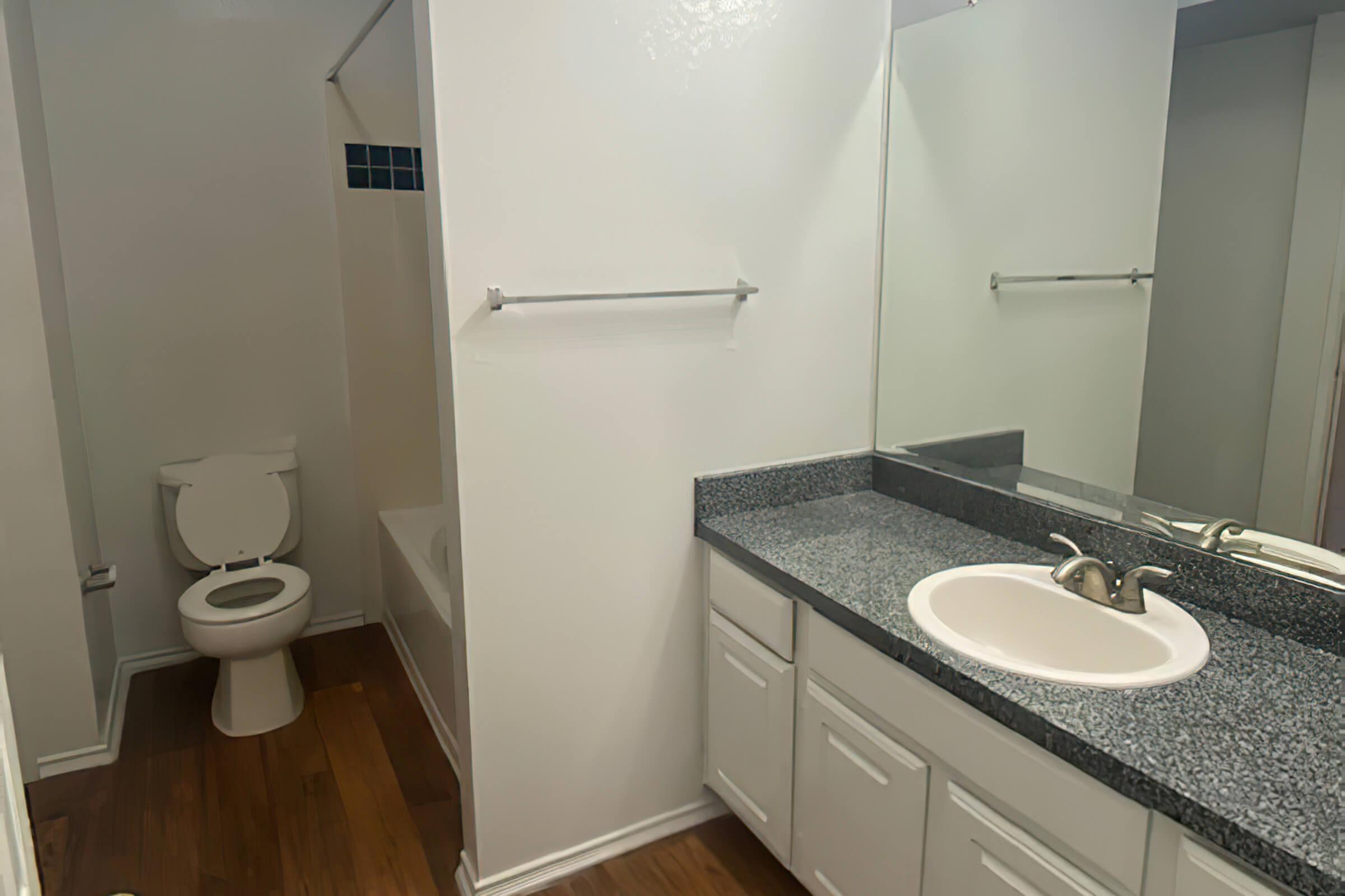 A clean, modern bathroom featuring a white bathtub with a shower, a toilet, a sink with a faucet on a granite countertop, and white cabinets. The walls are painted in a light color, and there is a large mirror above the sink. The floor is wooden, adding warmth to the space.