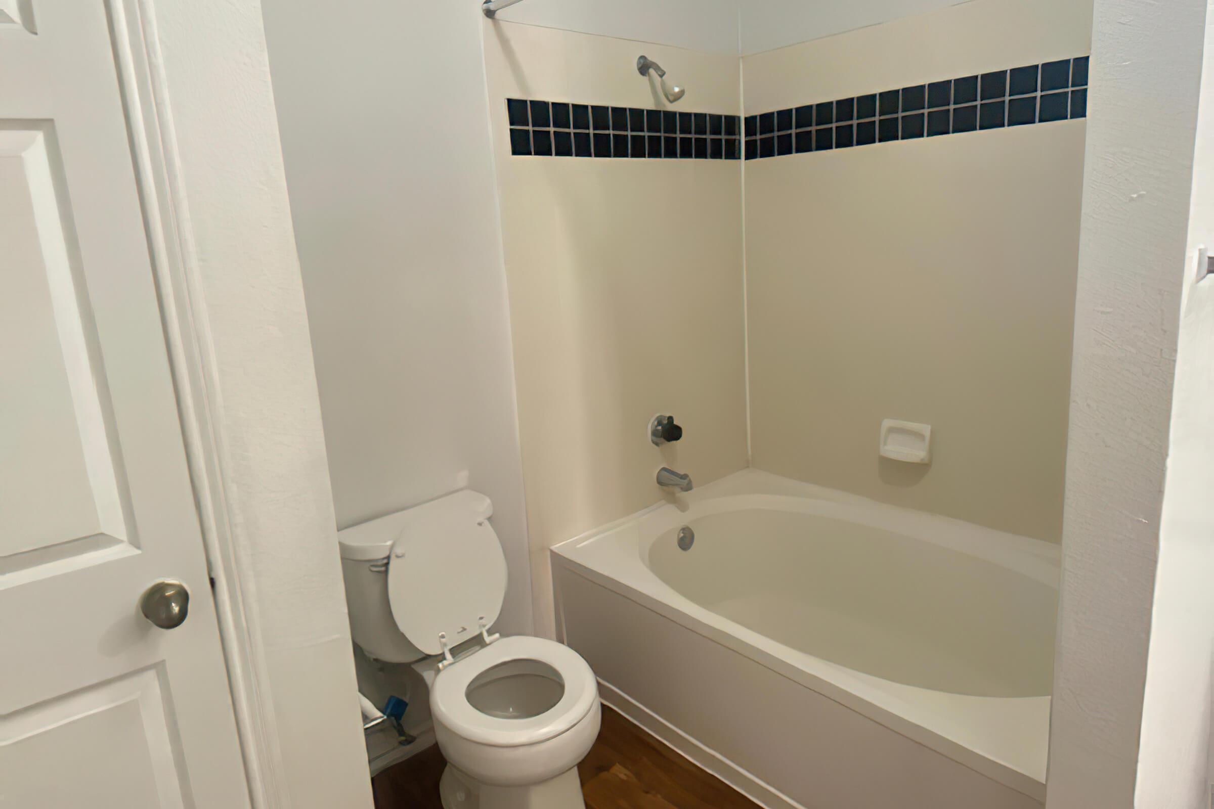 A small bathroom featuring a bathtub with a showerhead, a toilet, and a tiled section along the wall. The space has a light-colored wall and wooden flooring, with a single white door visible.