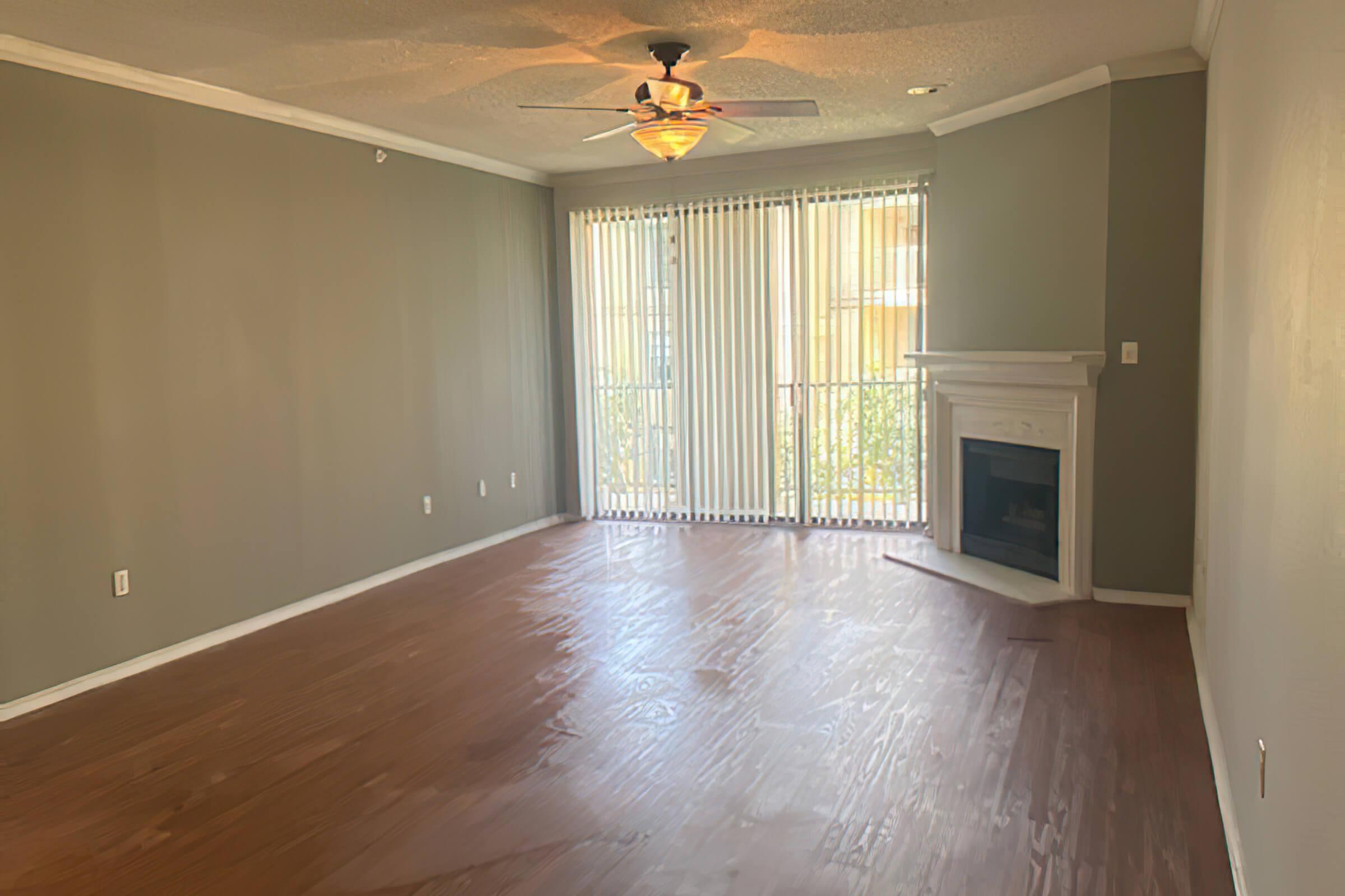 A spacious living room with light brown wooden floors, a ceiling fan, and a decorative fireplace. Large windows with vertical blinds allow natural light to fill the space. The walls are painted a soft green, and the atmosphere is inviting and bright.