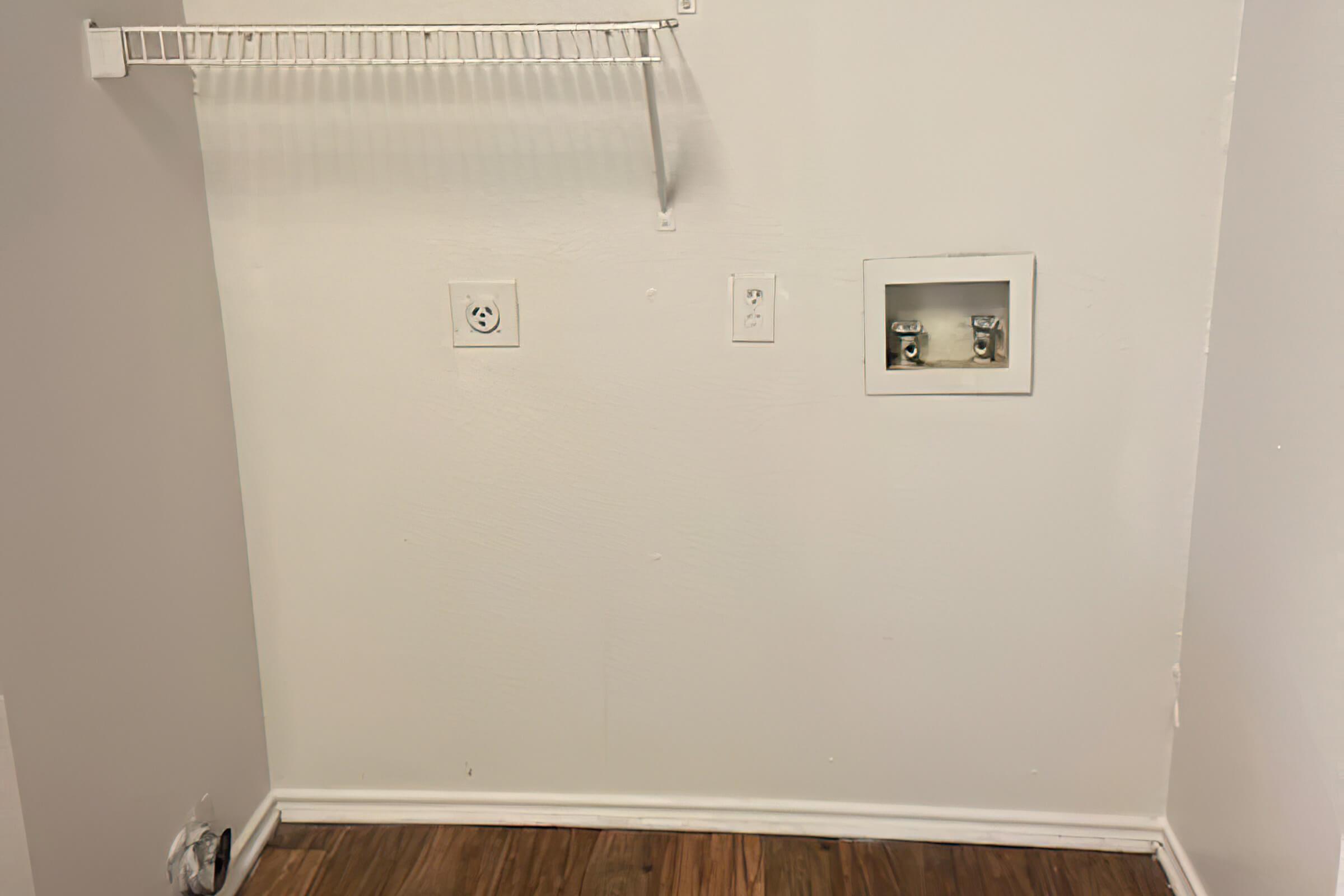 A plain wall with a white coat rack on the left, two electrical outlets and a utility box on the right. The floor is wood laminate, and the wall is painted white. The space appears empty and ready for use, possibly intended for a laundry area or storage.