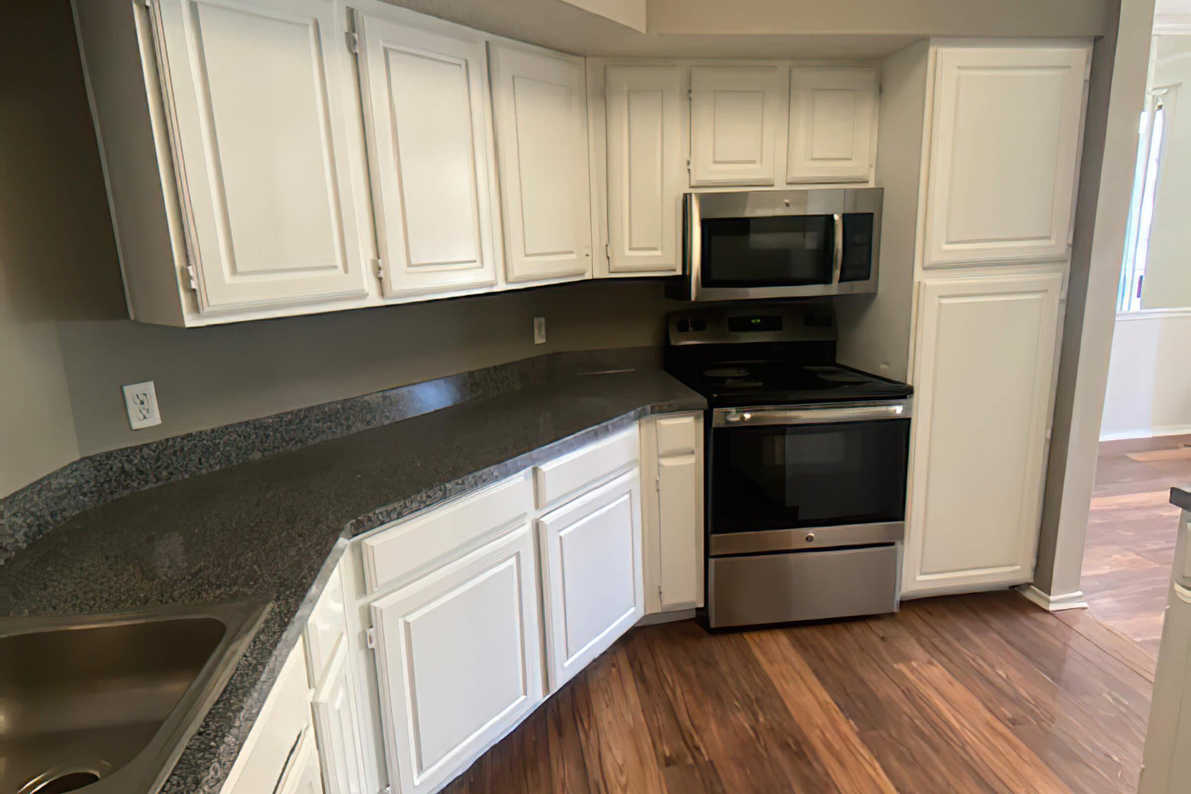 A modern kitchen featuring white cabinets, a stainless steel microwave, and an oven range. The countertops are dark granite, and there’s a stainless steel sink. The flooring is wooden, and there’s natural light coming through a nearby window. The overall design is clean and contemporary.