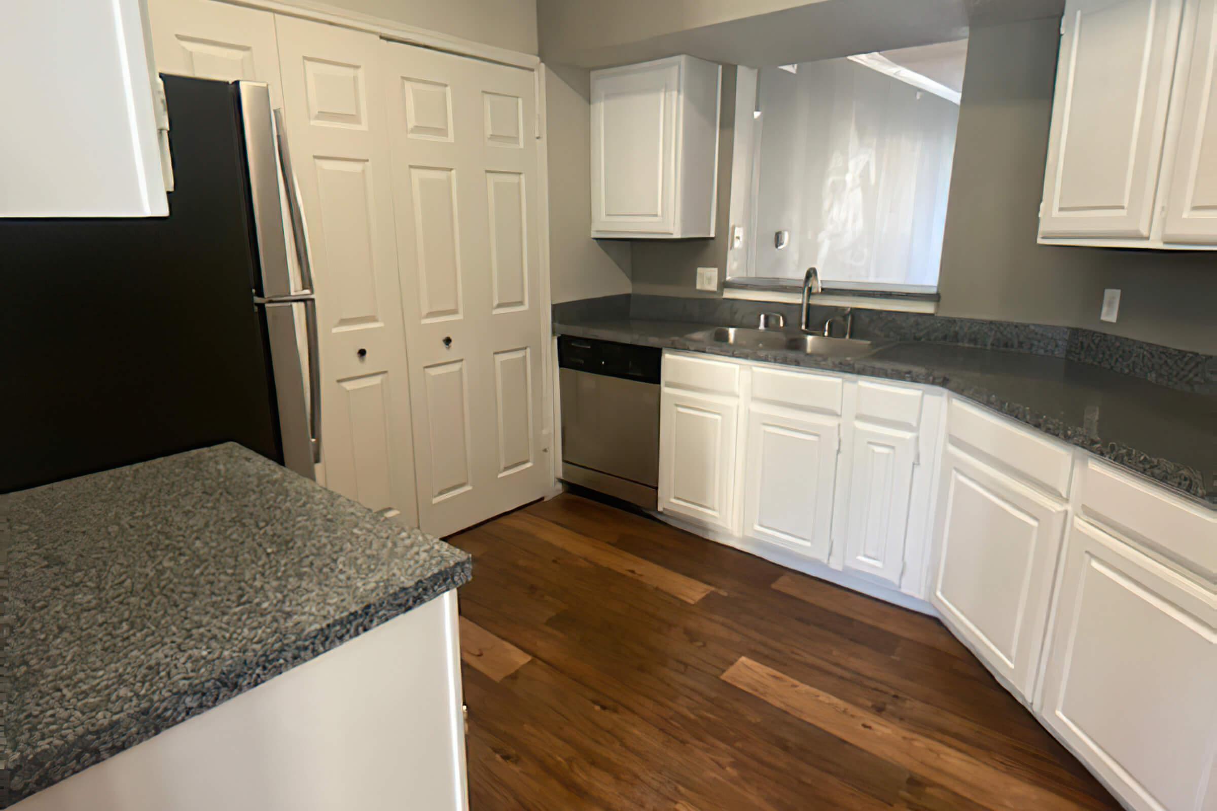 Modern kitchen featuring light gray walls, white cabinetry, and dark granite countertops. Includes stainless steel appliances like a refrigerator and dishwasher, along with a spacious sink. The floor is made of wood, creating a warm atmosphere. Bright lighting enhances the overall openness of the space.