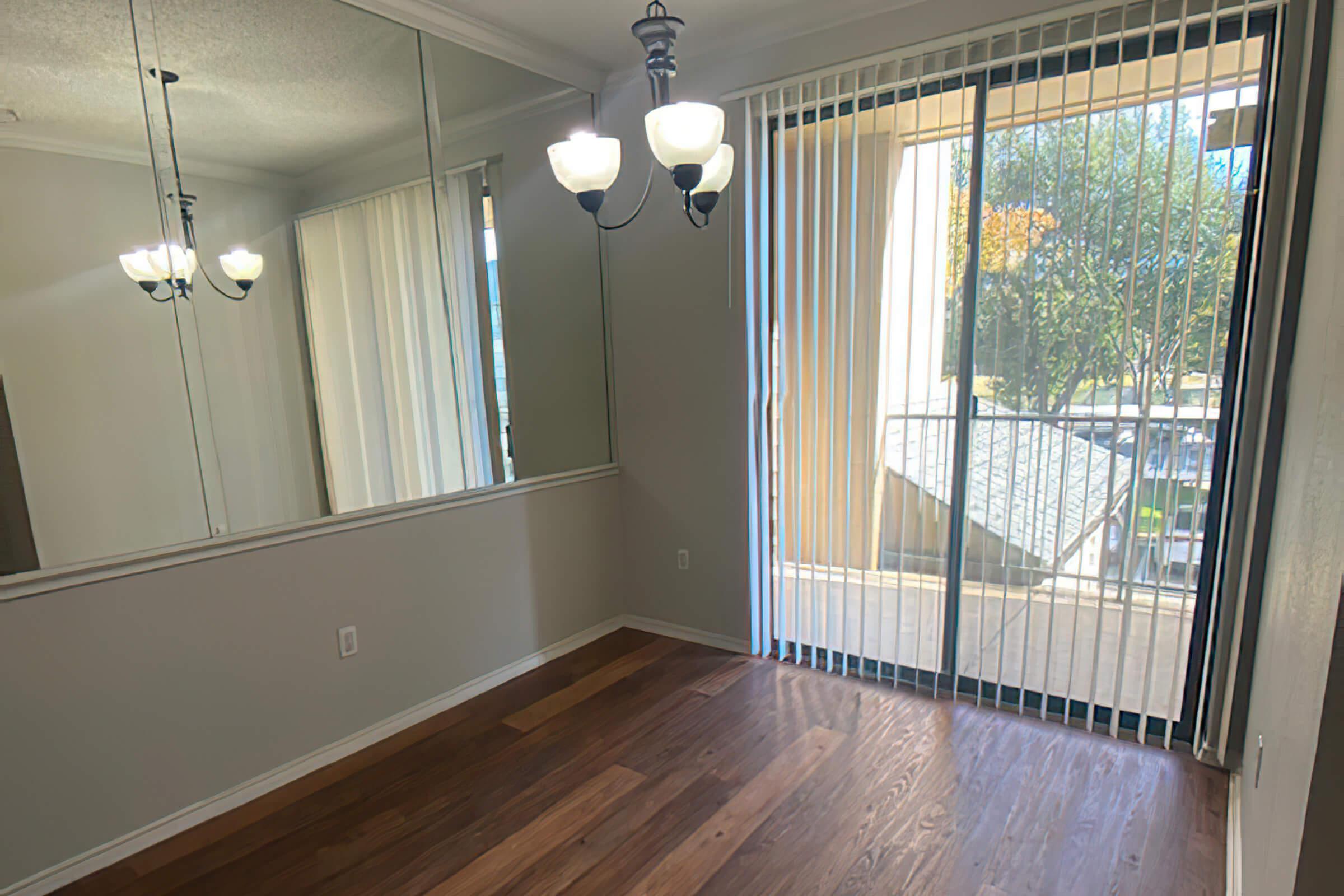 A well-lit room with hardwood floor and large sliding glass door leading to a balcony. The walls are light-colored, and there is a mirror on one wall reflecting the space. A modern chandelier with three lights hangs from the ceiling, adding to the airy atmosphere.