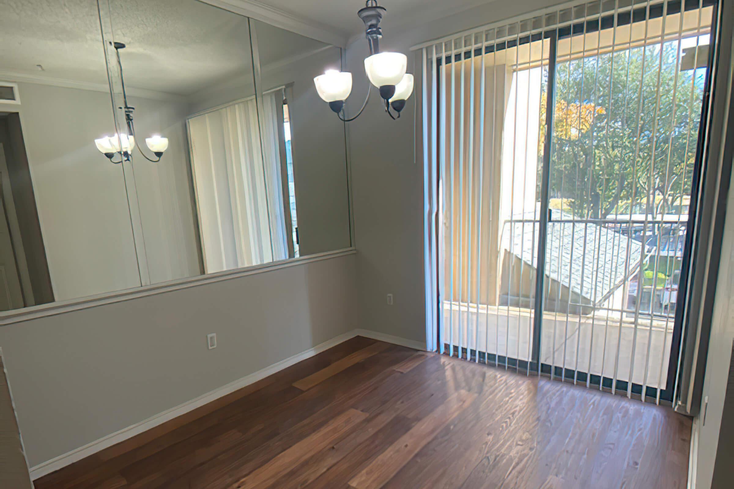 A bright room with hardwood flooring, featuring large windows with vertical blinds and a sliding glass door leading to a balcony. The wall opposite the windows has mirrors and a modern chandelier with three light bulbs, creating an open and airy atmosphere.