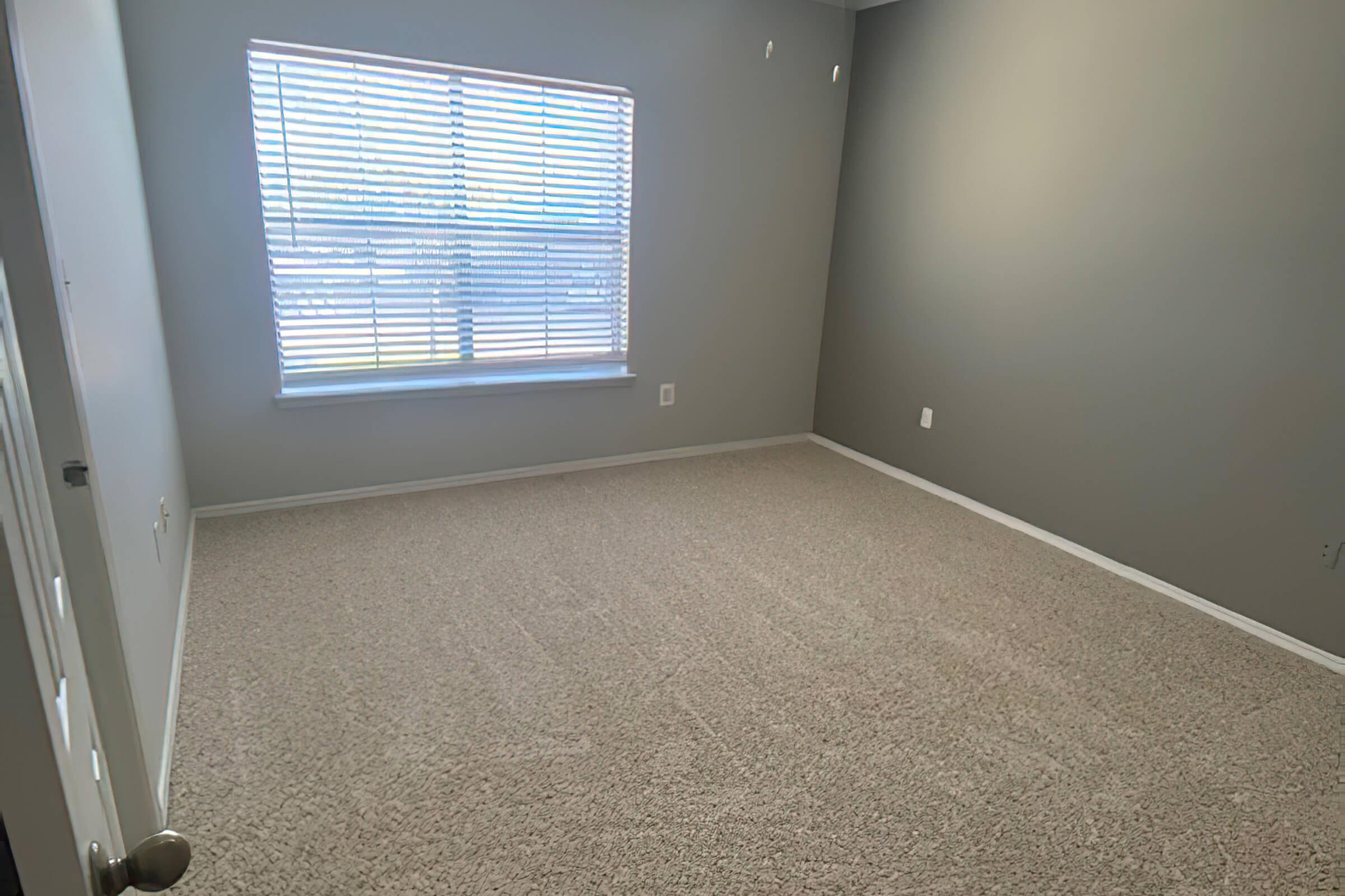 A vacant room with light gray walls and beige carpet. A window with white blinds allows natural light into the space, illuminating the bare floor. There are no furnishings or decorations, creating an open and minimalist atmosphere. A door is visible on the left side of the image.