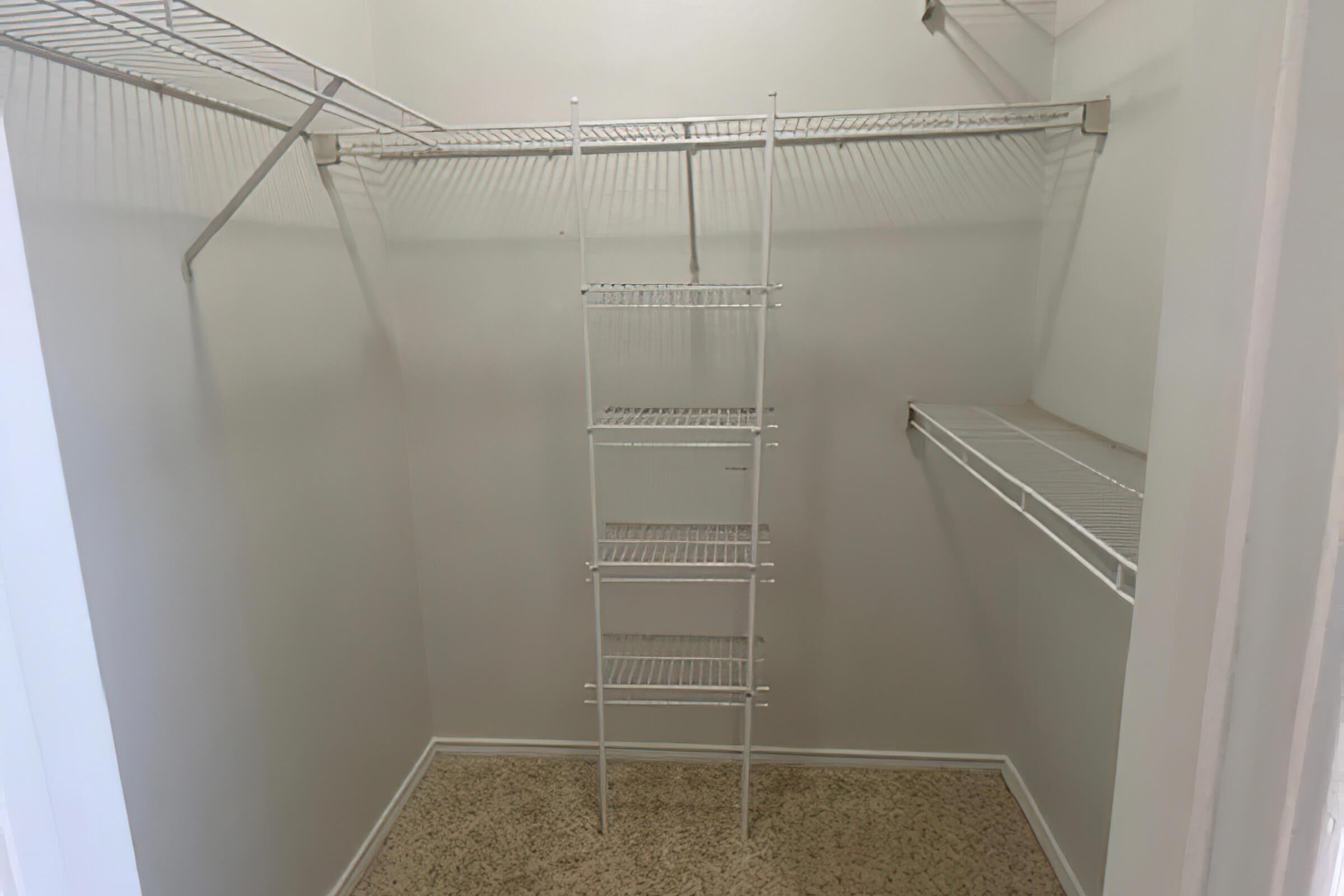 A clean and empty walk-in closet featuring white wire shelving and racks mounted on the walls. The floor is carpeted, and there is a metal shelving unit in the center for additional storage. The walls are painted a light color, creating a bright and spacious atmosphere.