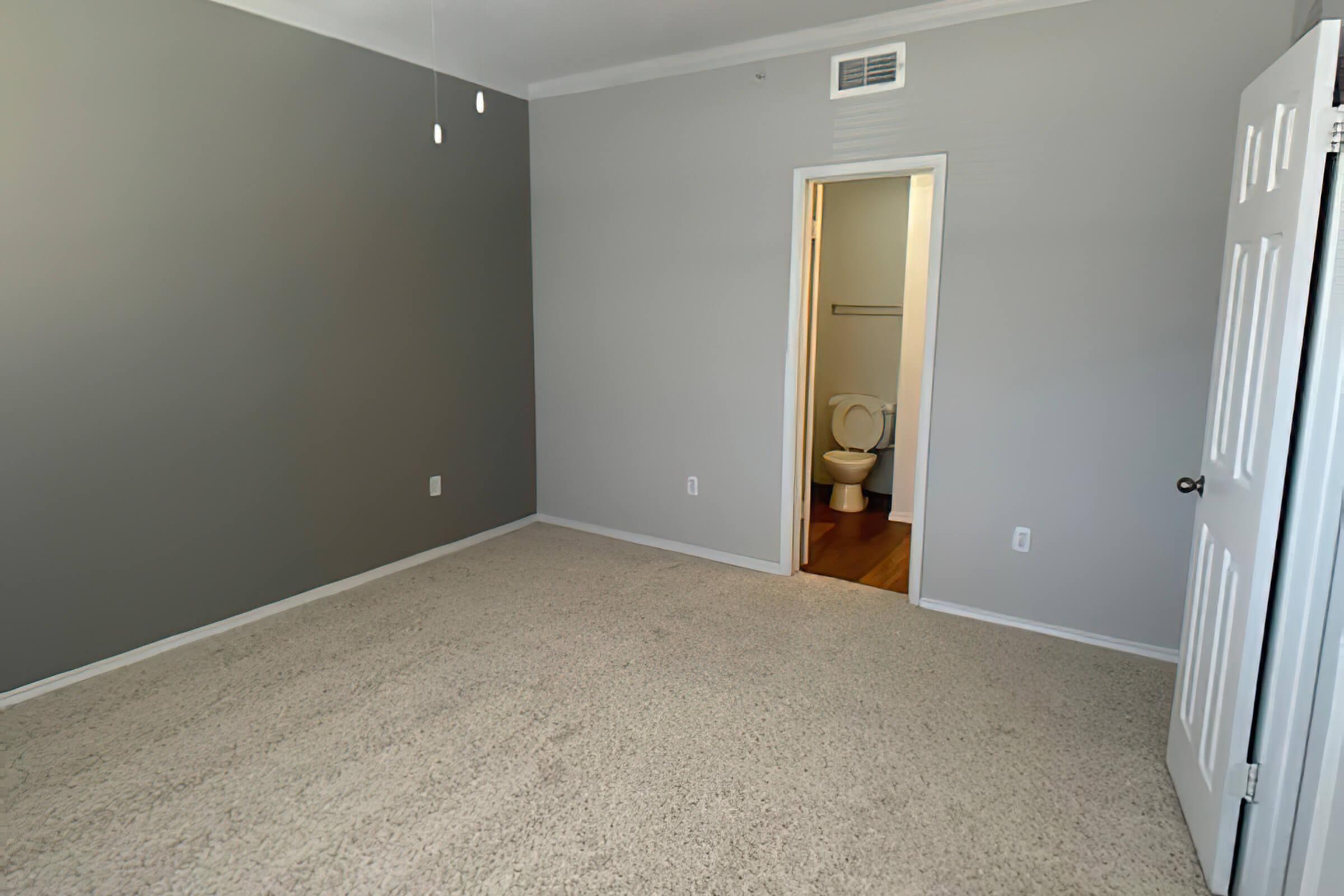 A spacious, empty room with light gray walls and beige carpet. A door leads to a bathroom visible in the background, featuring a toilet and light-colored flooring. The room has a ceiling fan with two pull strings and is well-lit by natural light.