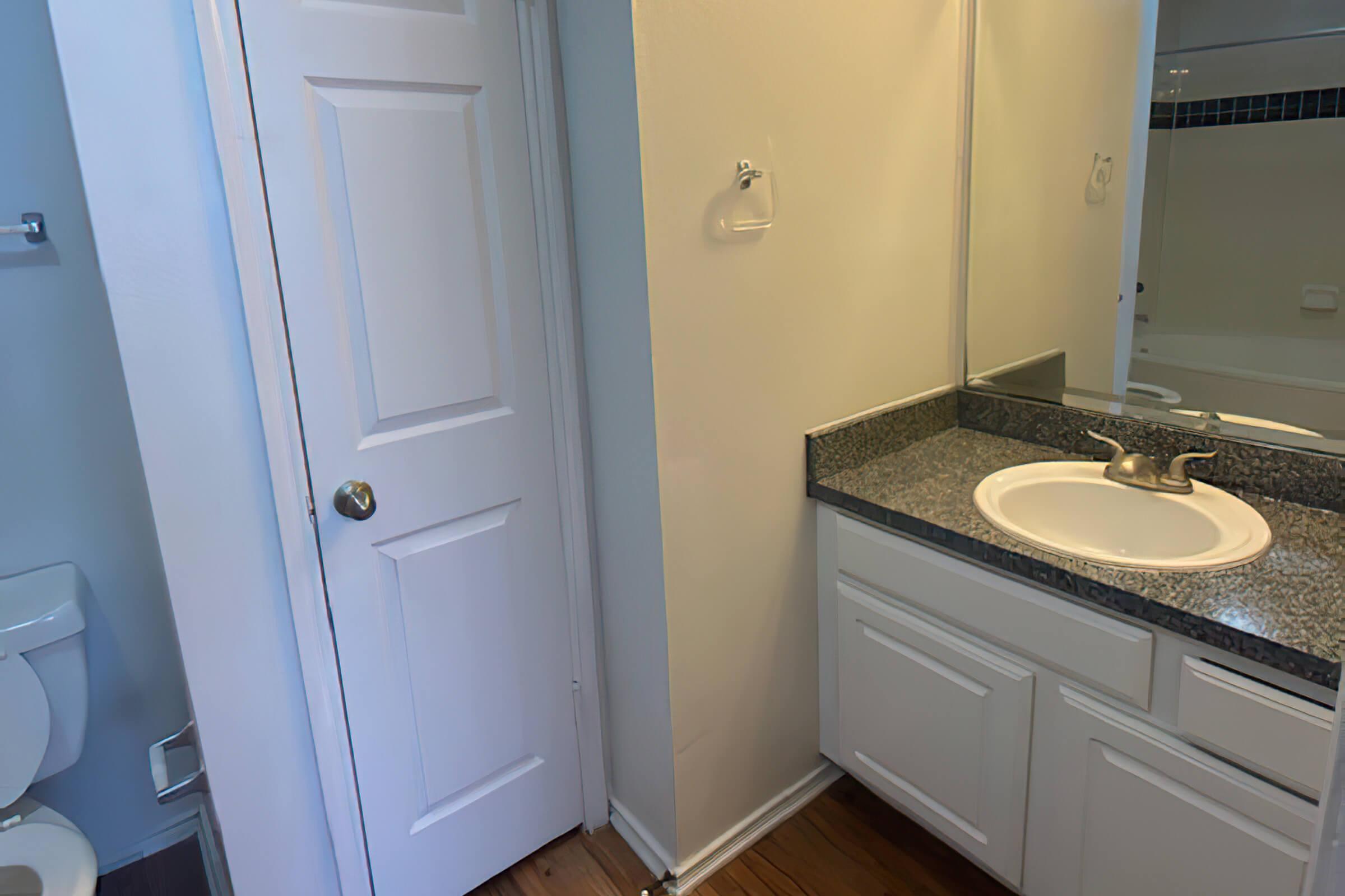 A bathroom view featuring a white door leading to a toilet area on the left, a granite countertop with a sink in the center, and a mirror above the sink. The walls are painted light colors, and there is a glimpse of a bathtub in the background. The flooring is wooden.