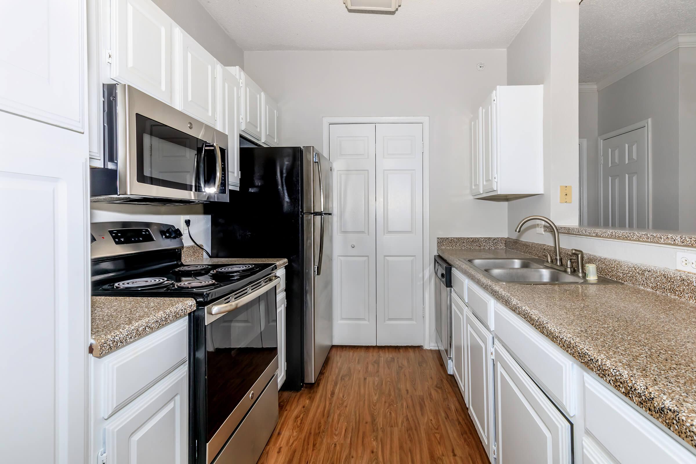 A modern kitchen featuring white cabinets, stainless steel appliances, including a microwave and refrigerator, a black oven range, granite countertops, and a single basin sink. The room has light-colored walls and wooden flooring, creating a bright and inviting atmosphere.