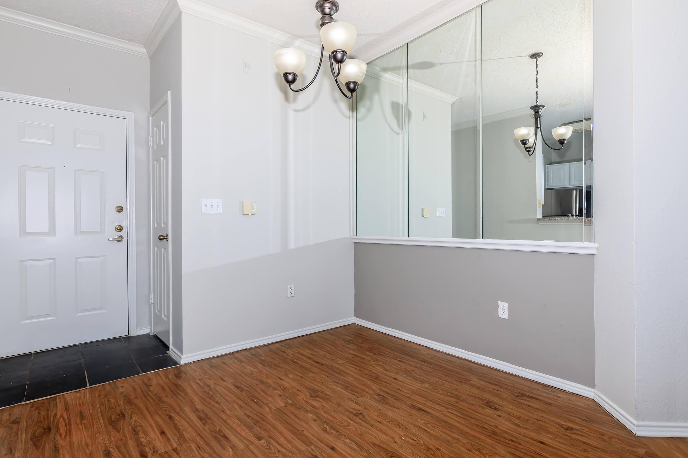 A small entryway featuring a light-colored wall, a mirrored section, and a hardwood floor. The area includes a doorway with a six-panel door, a wall-mounted light fixture with three bulbs, and a corner leading to a kitchen space visible in the background.