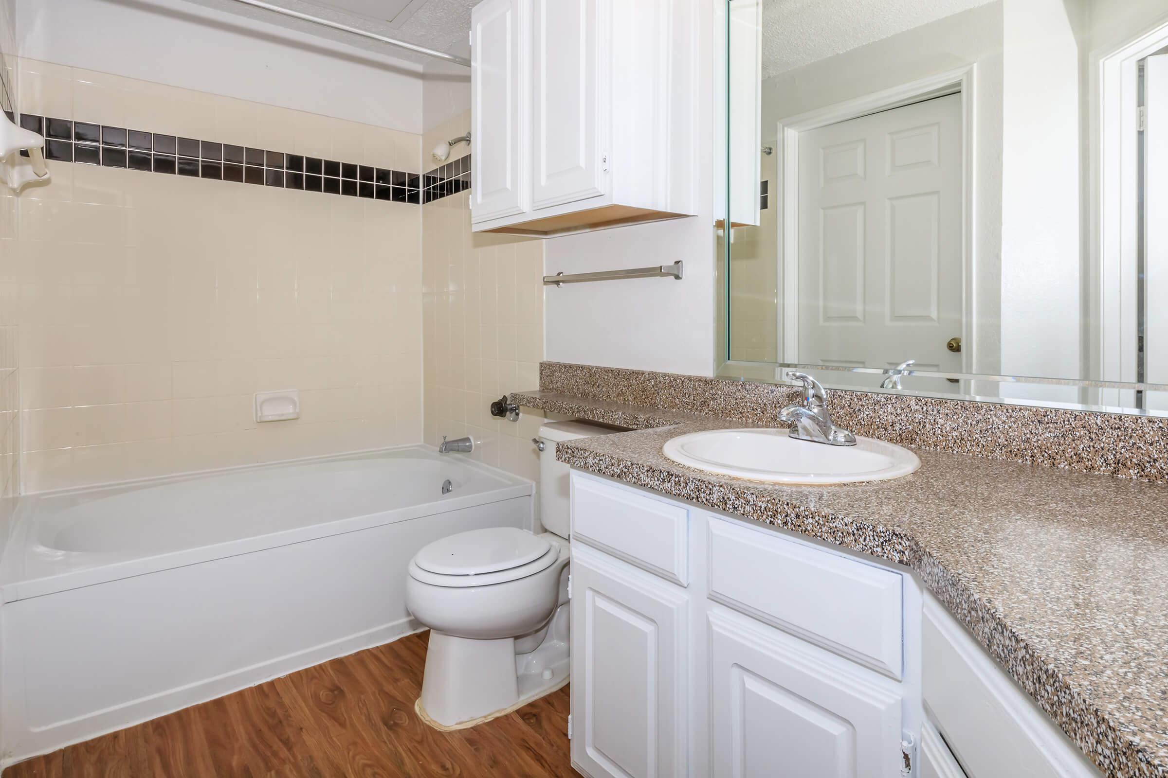 A clean bathroom featuring a white bathtub with a shower, a granite countertop with a circular sink, and white cabinetry. The walls are beige with a black tile accent, and there is a mirror above the sink. The floor is covered in wood-like vinyl. A door leads to another room.