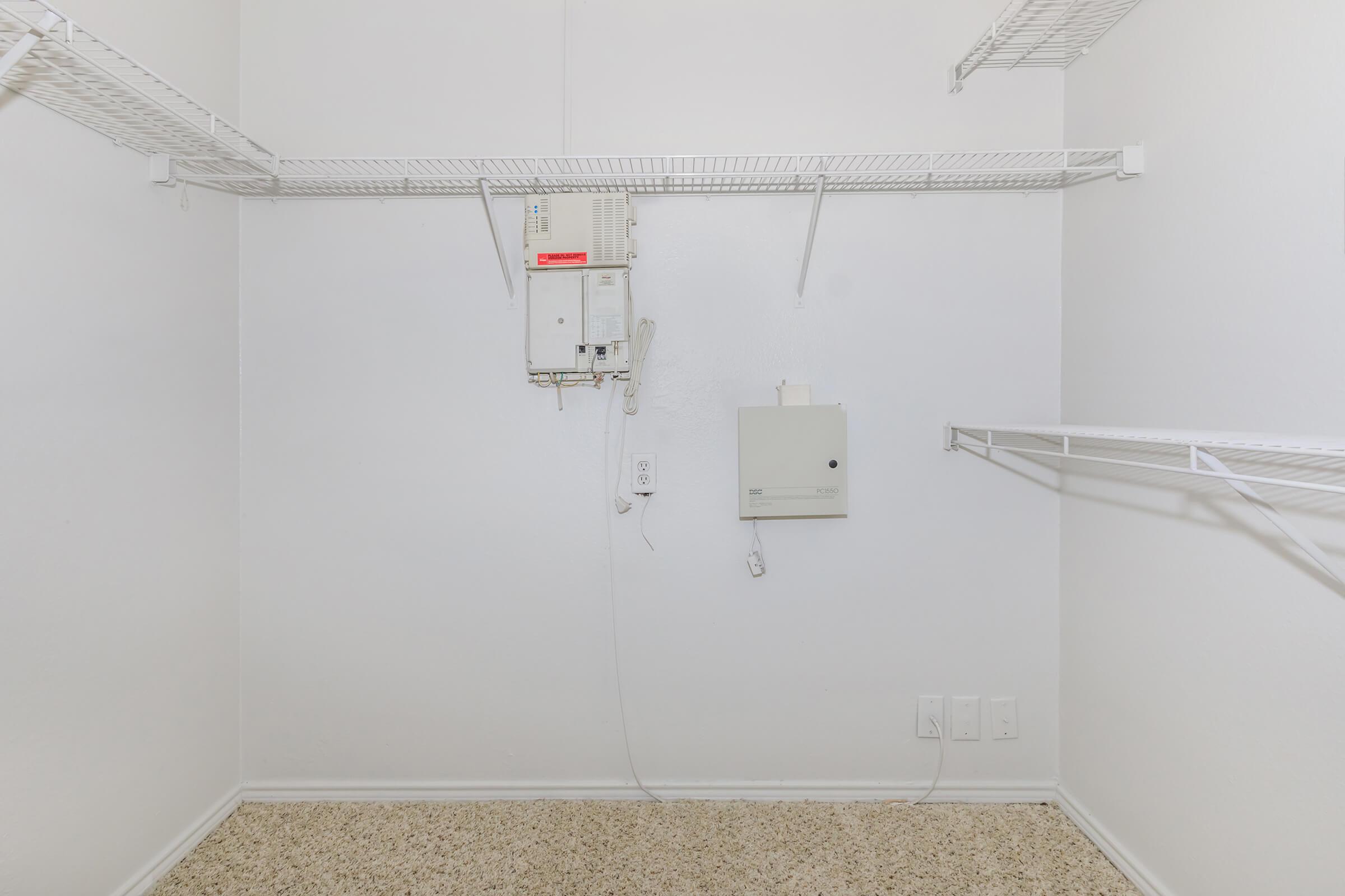 Empty closet with white walls and light beige carpeting. There are wire shelves on the left and right sides, and two communication panels mounted on the wall. The space is uncluttered and well-lit, suggesting a neat and organized environment.