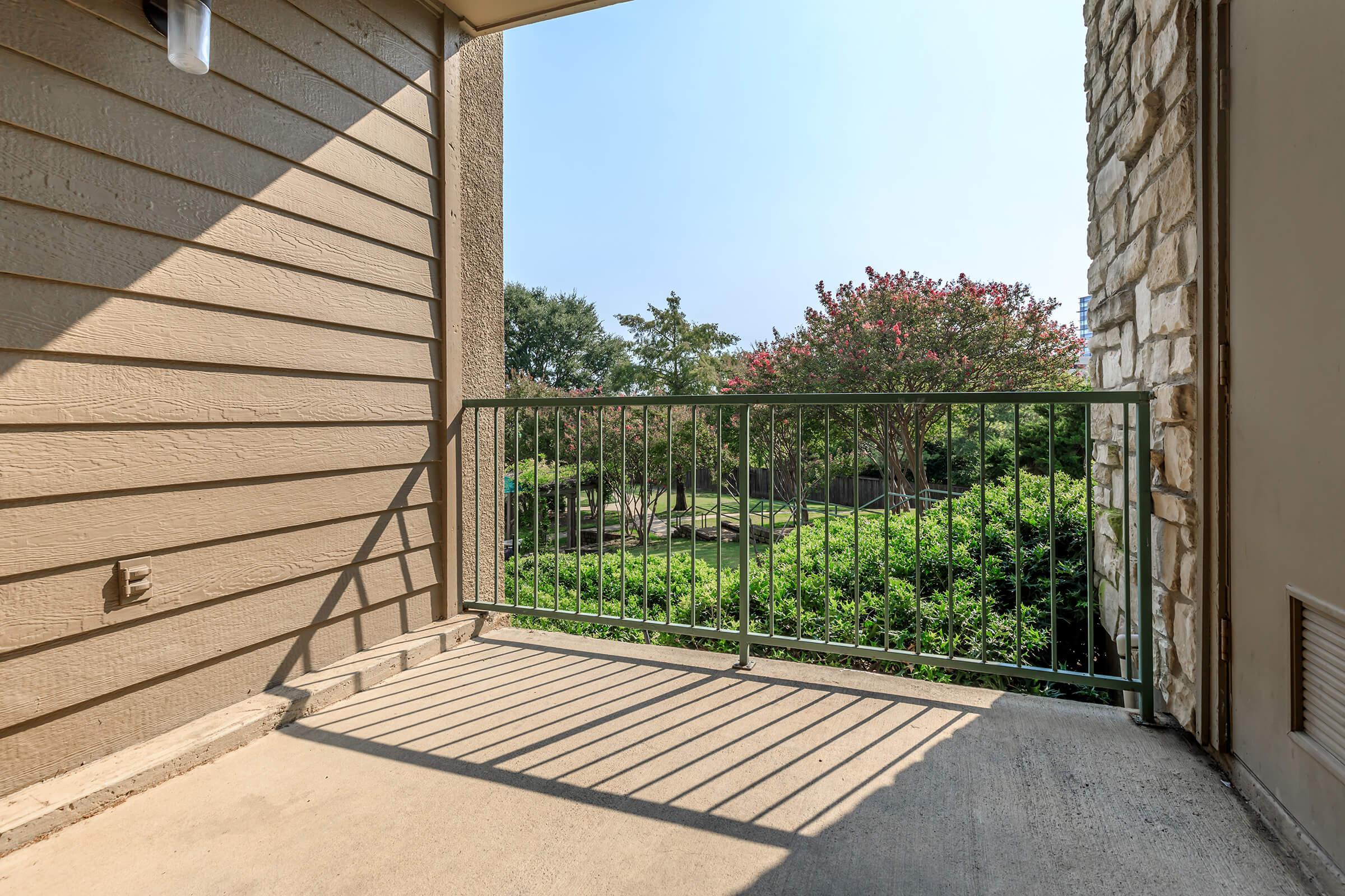 A balcony view featuring a green railing with shadows cast on the floor. In the background, there are lush green bushes and trees, with bright blue sky above. The scene conveys a peaceful outdoor space, perfect for relaxation.