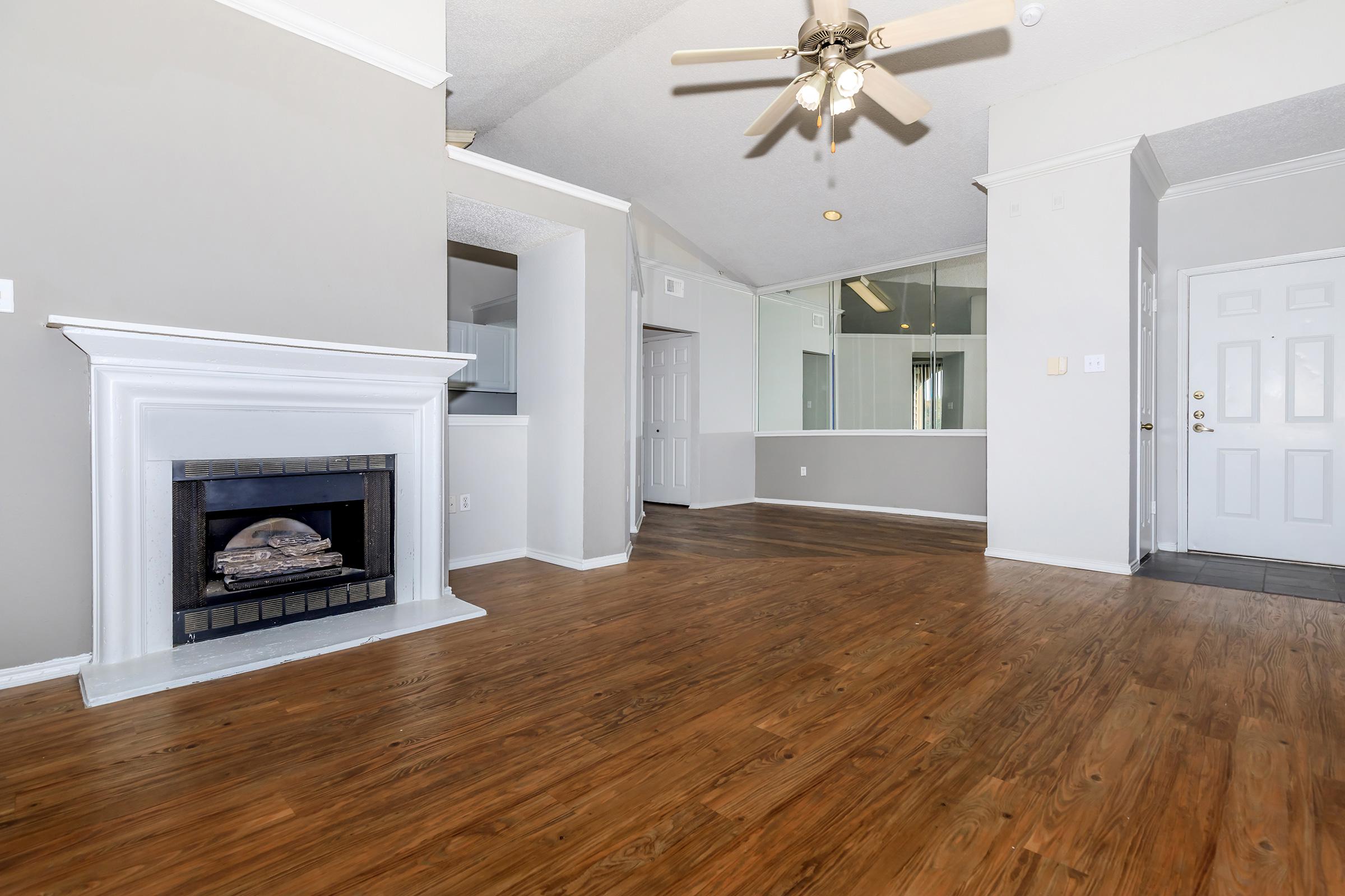 Spacious interior of a living room featuring a fireplace, light-colored walls, and hardwood flooring. A ceiling fan is visible, and there is a mirrored wall that adds depth to the space. The entrance door is located in the corner, and natural light is coming through the windows.