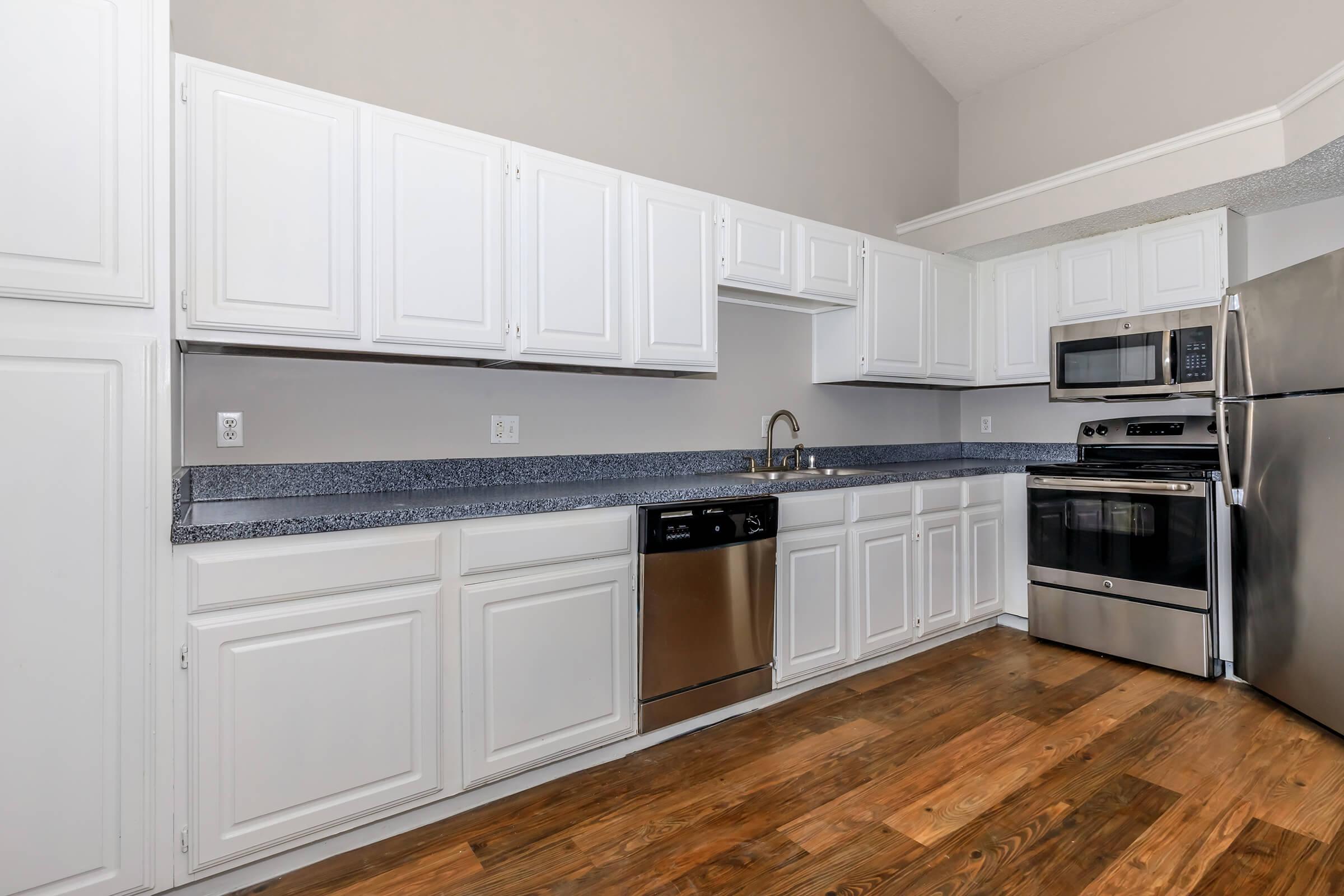 Modern kitchen featuring white cabinetry, granite countertops, stainless steel appliances including a dishwasher, oven, and microwave. The space has a warm wooden floor and light gray walls, creating a bright and inviting atmosphere.