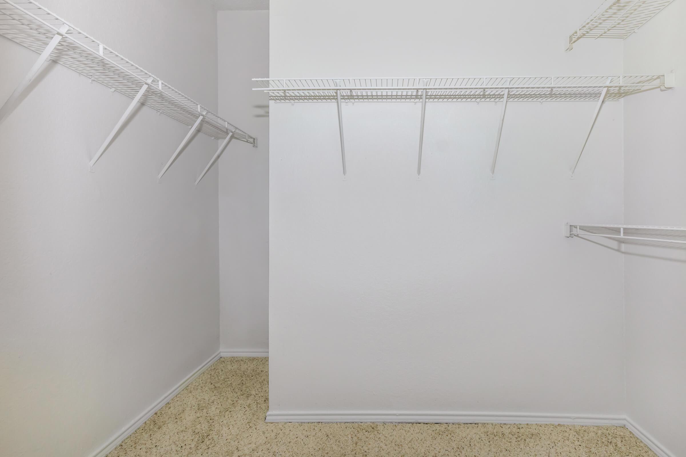 Empty walk-in closet with white walls and a light-colored carpet. The closet features wire shelving on two sides, with several hanging rods for clothing. The space is well-lit and primarily unadorned, creating a minimalistic and spacious appearance.