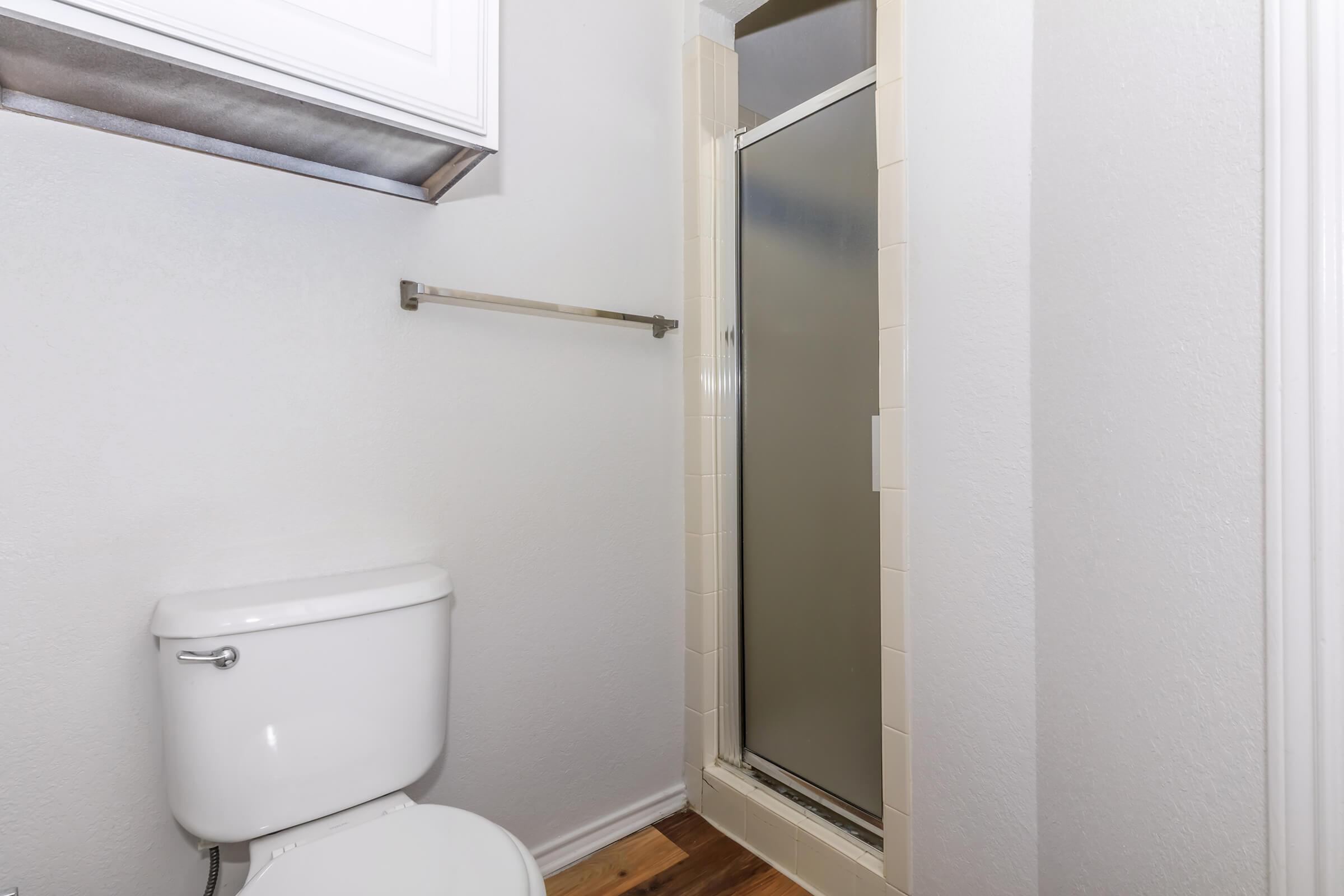 A small bathroom featuring a white toilet and a glass shower enclosure. The walls are painted light gray, and there is a metal towel bar mounted on the wall. The flooring is wooden, contributing to a clean and simple design.