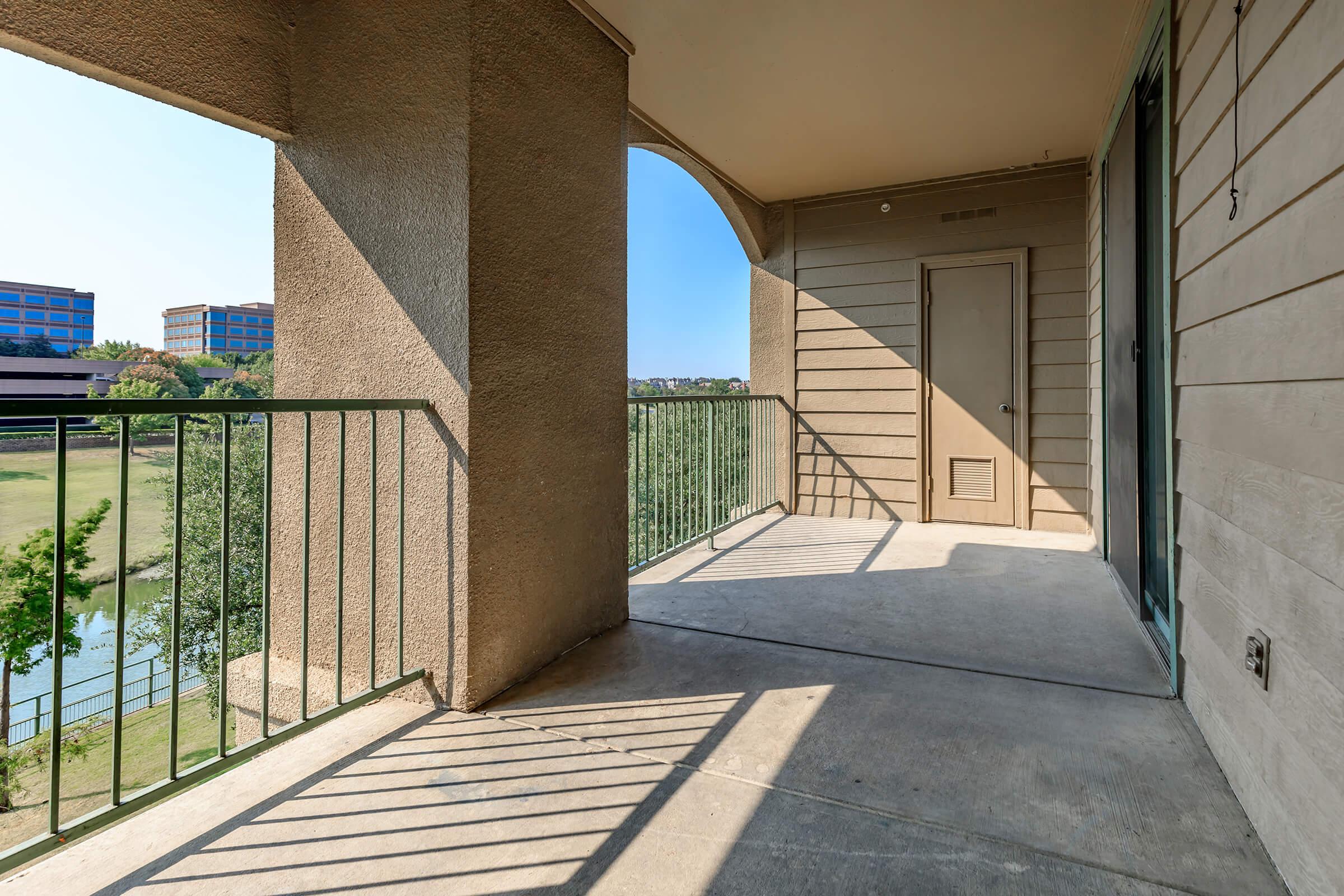 Balcony view featuring a concrete floor, green railing, and sunlight casting shadows. The background shows greenery and buildings in the distance. The area appears spacious and well-lit, creating a serene outdoor space.