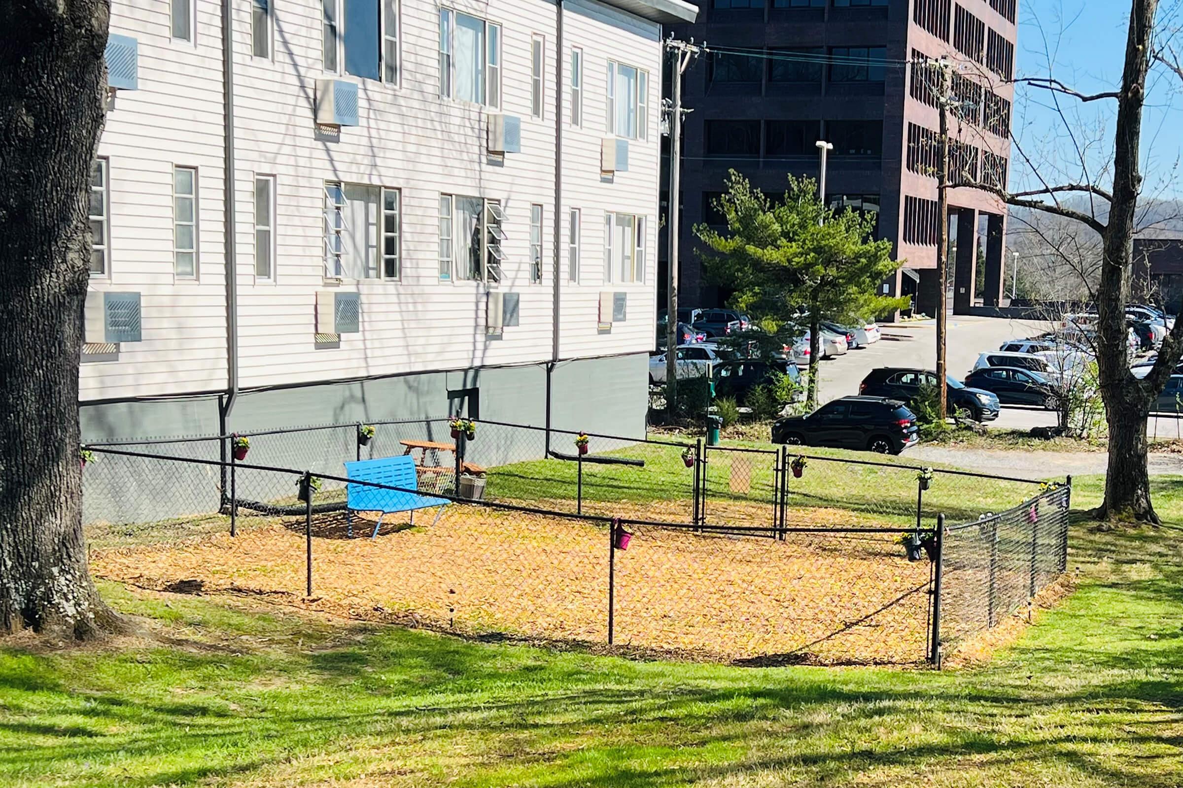 A fenced dog park featuring a sandy ground and a small blue bench. The park is surrounded by grassy areas and is located near two multi-story buildings. There are trees nearby, and cars are parked in the background.