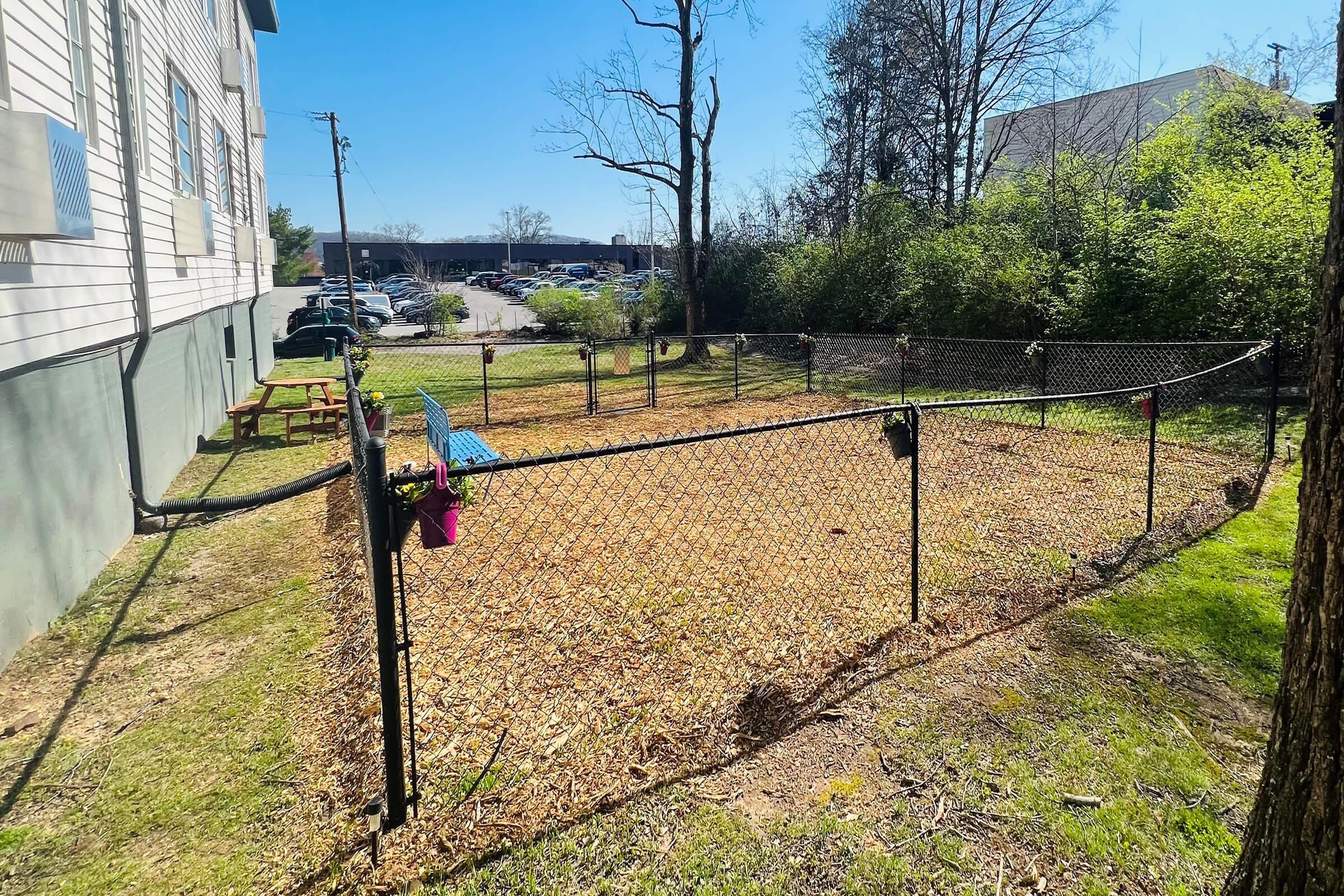A fenced outdoor area with a dirt ground, surrounded by trees and greenery. There is a picnic table and a blue bench inside the enclosure, with a few plastic cups hanging on the fence. The background features a parking lot and commercial buildings under a clear blue sky.