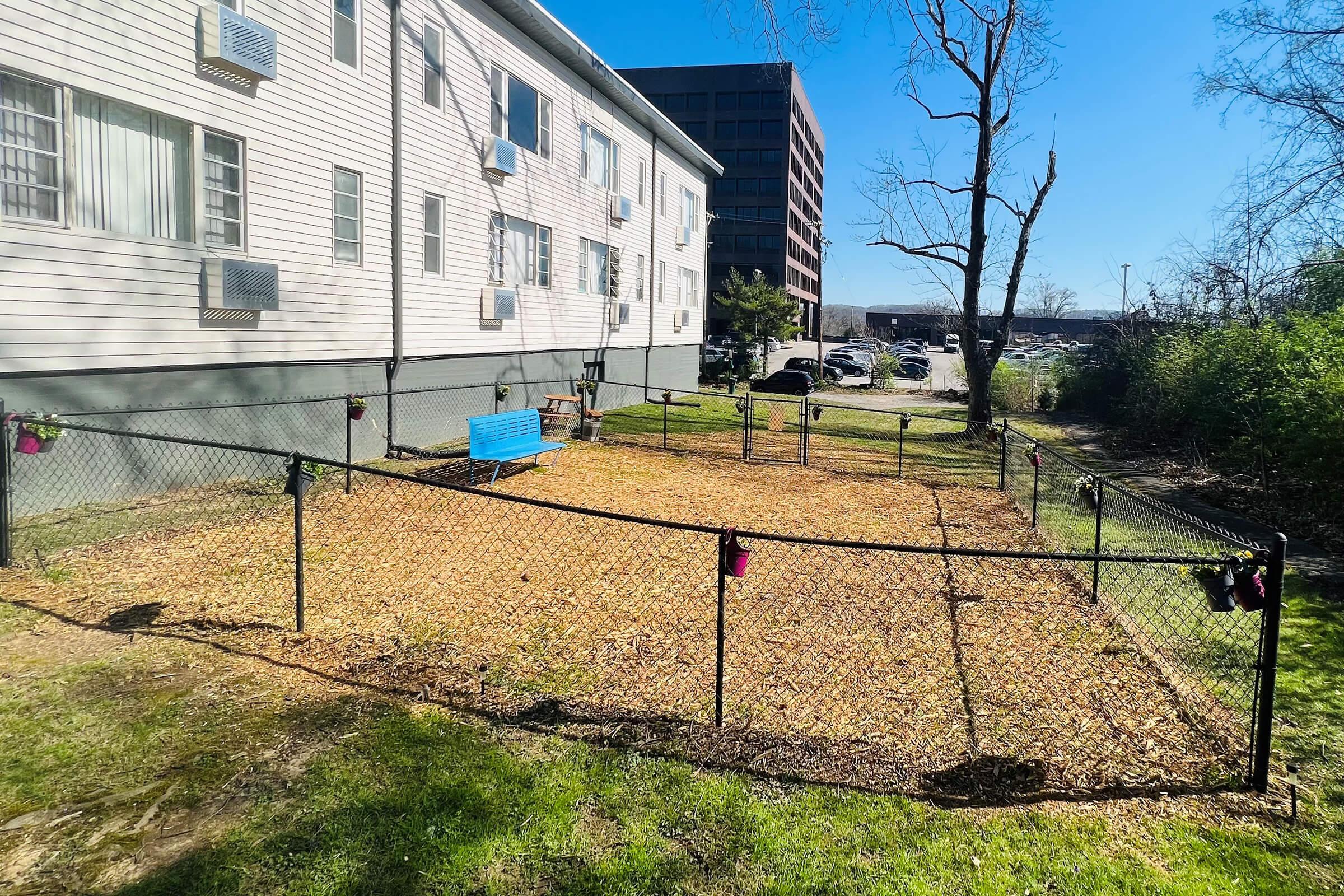 A fenced outdoor area with wood chips, featuring a blue bench and hanging pots, adjacent to a building and parking lot. The setting is sunny with clear skies and trees in the background.
