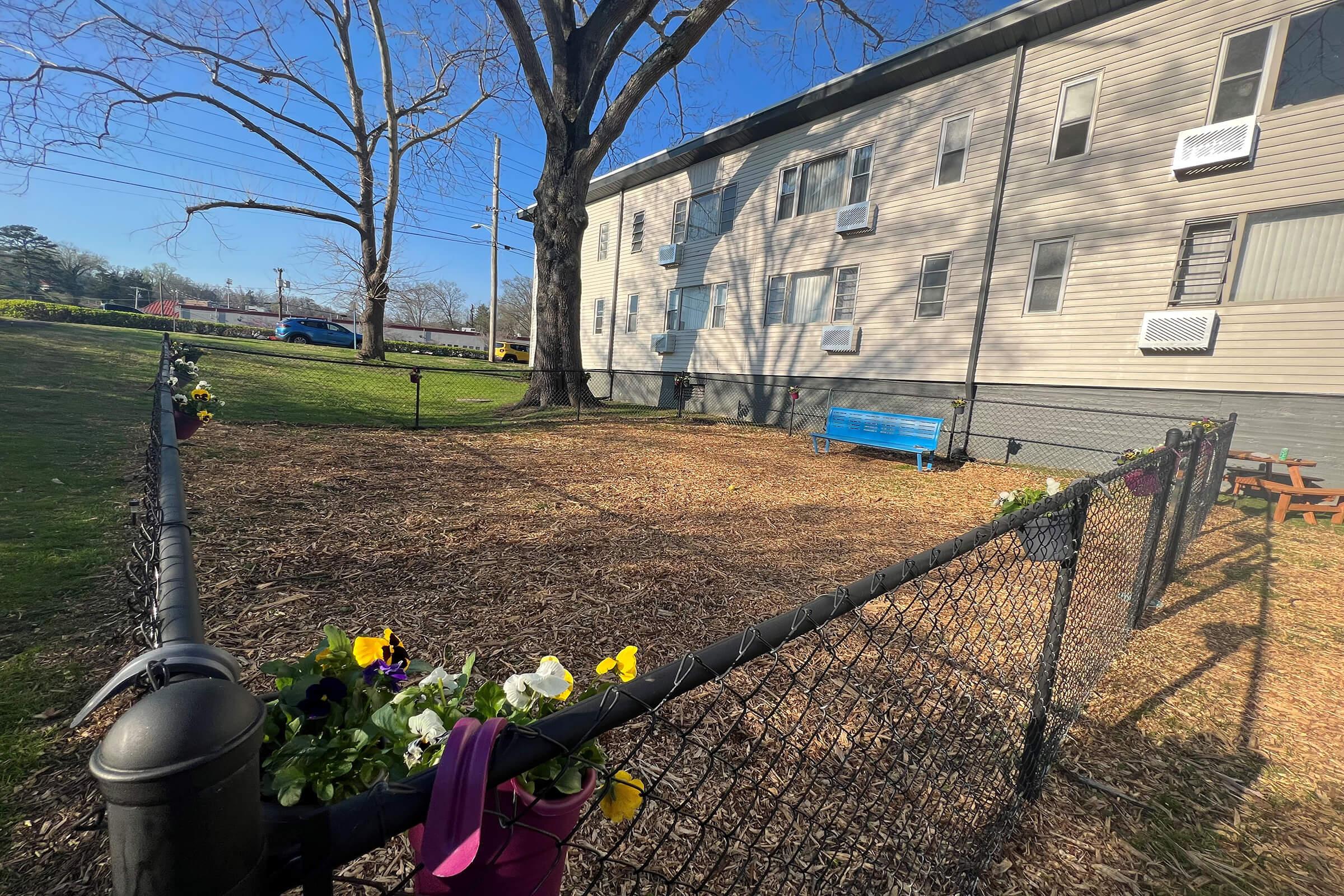 A fenced area with mulch and flowers along the fence, situated next to a multi-story building. A blue bench is visible inside the enclosure, and a picnic table is positioned nearby. Trees and clear blue skies create a peaceful outdoor environment.