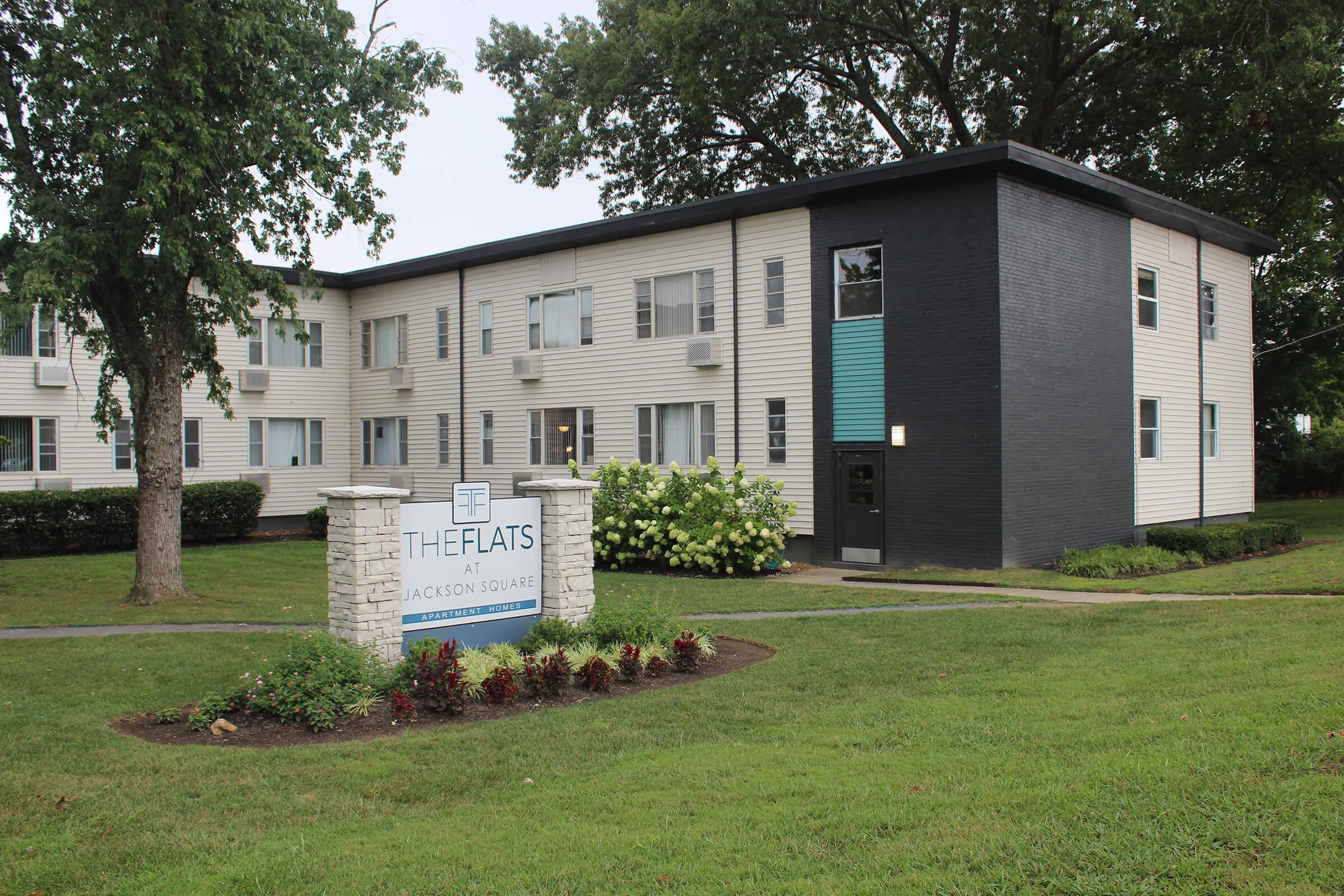 A modern apartment building with a landscaped lawn, featuring a prominent sign that reads "The Flats at Jackson Square." The building has a mix of light and dark siding, with several windows, and is surrounded by bushes and trees, indicating a well-maintained residential area.