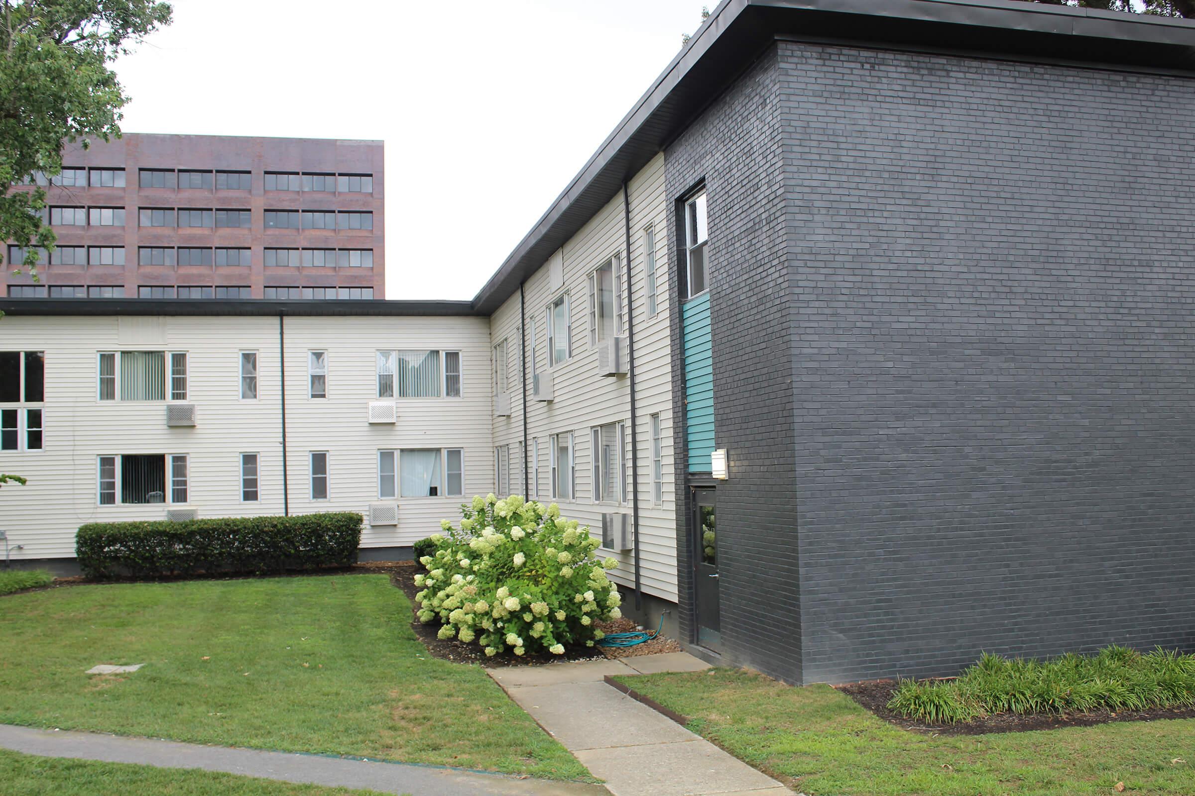A modern multi-unit residential building with a combination of light-colored siding and dark brick. In the foreground, there's a well-maintained grassy area with a large flowering bush. The background features a taller building. The scene is set on a cloudy day, giving a calm, subdued atmosphere.