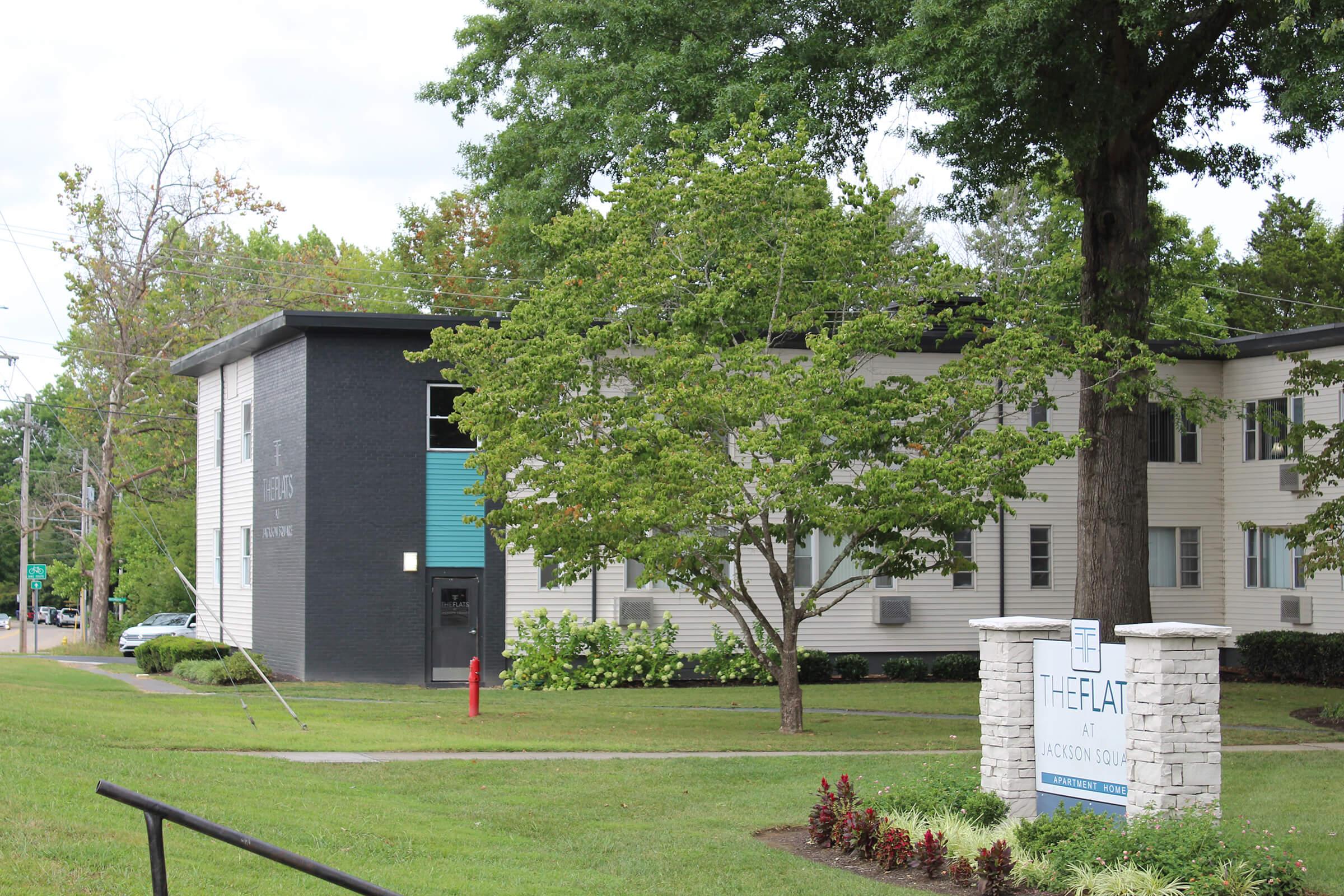 A modern apartment building featuring a combination of dark and light siding, surrounded by green grass and trees. A sign in front reads "The Flat at Action Sol," indicating the name of the apartments. The scene captures a well-maintained exterior with a calm, residential atmosphere.