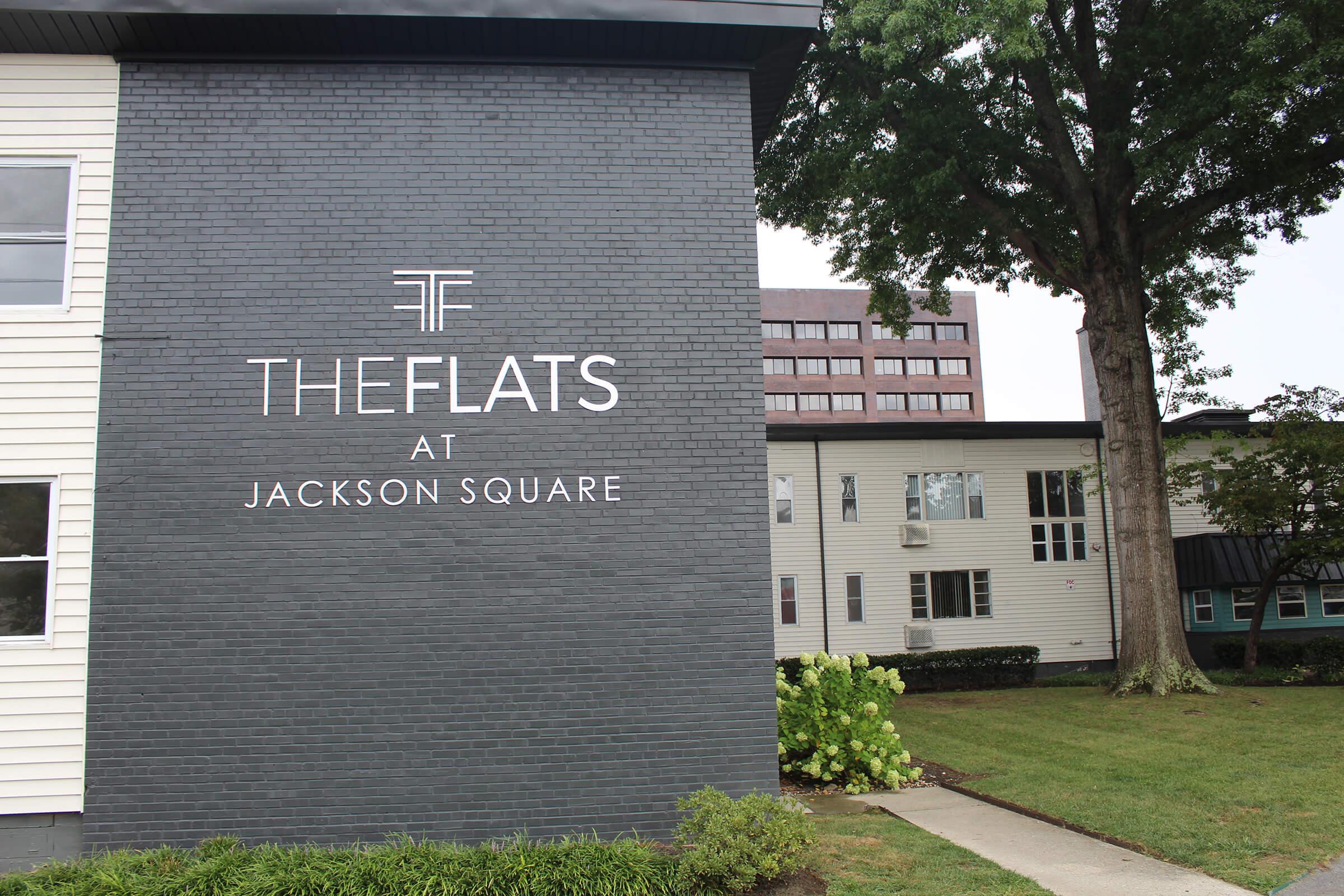 Signage for "The Flats at Jackson Square" on a dark brick wall, with modern font. Surrounding area features greenery and residential buildings. The setting is urban, with a mix of architectural styles.