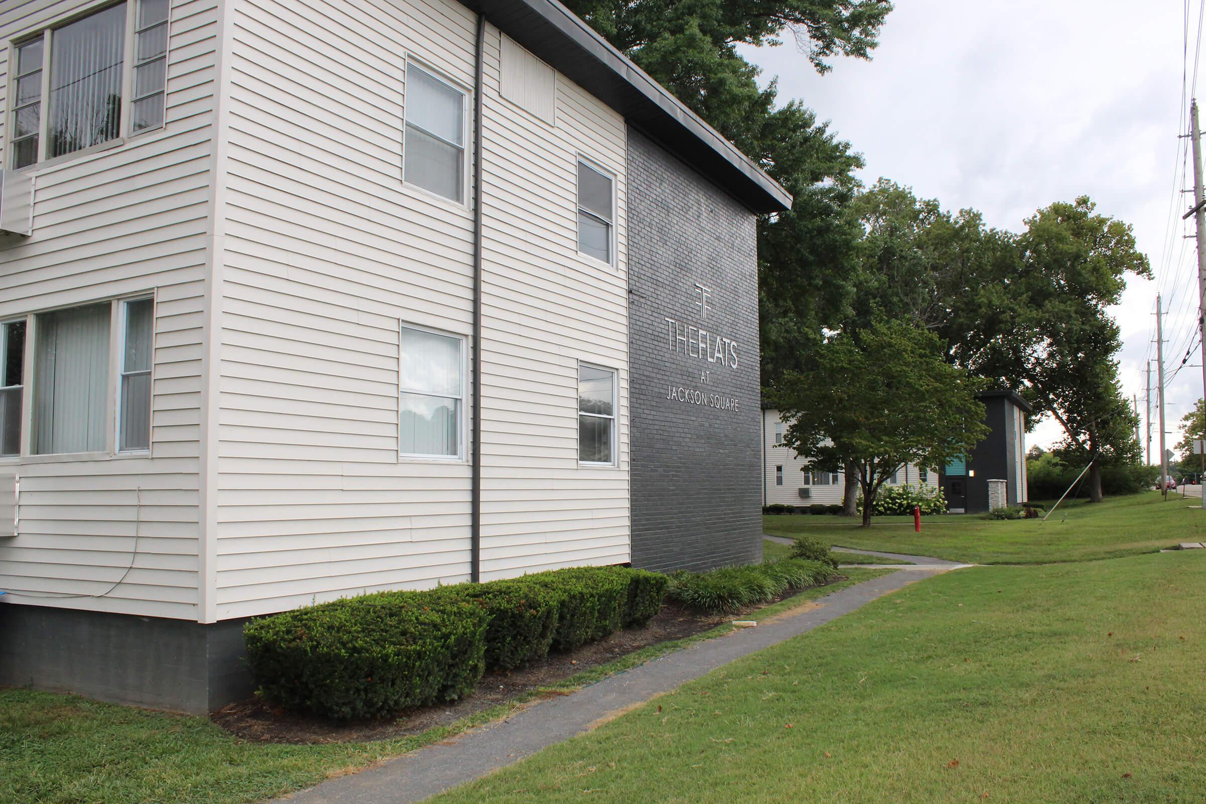 A view of a residential building featuring two distinct sections: one side with a white wooden exterior and large windows, and the other side with a dark brick facade displaying the words "The Flats." The surrounding area has well-maintained grass and shrubs, with trees in the background.