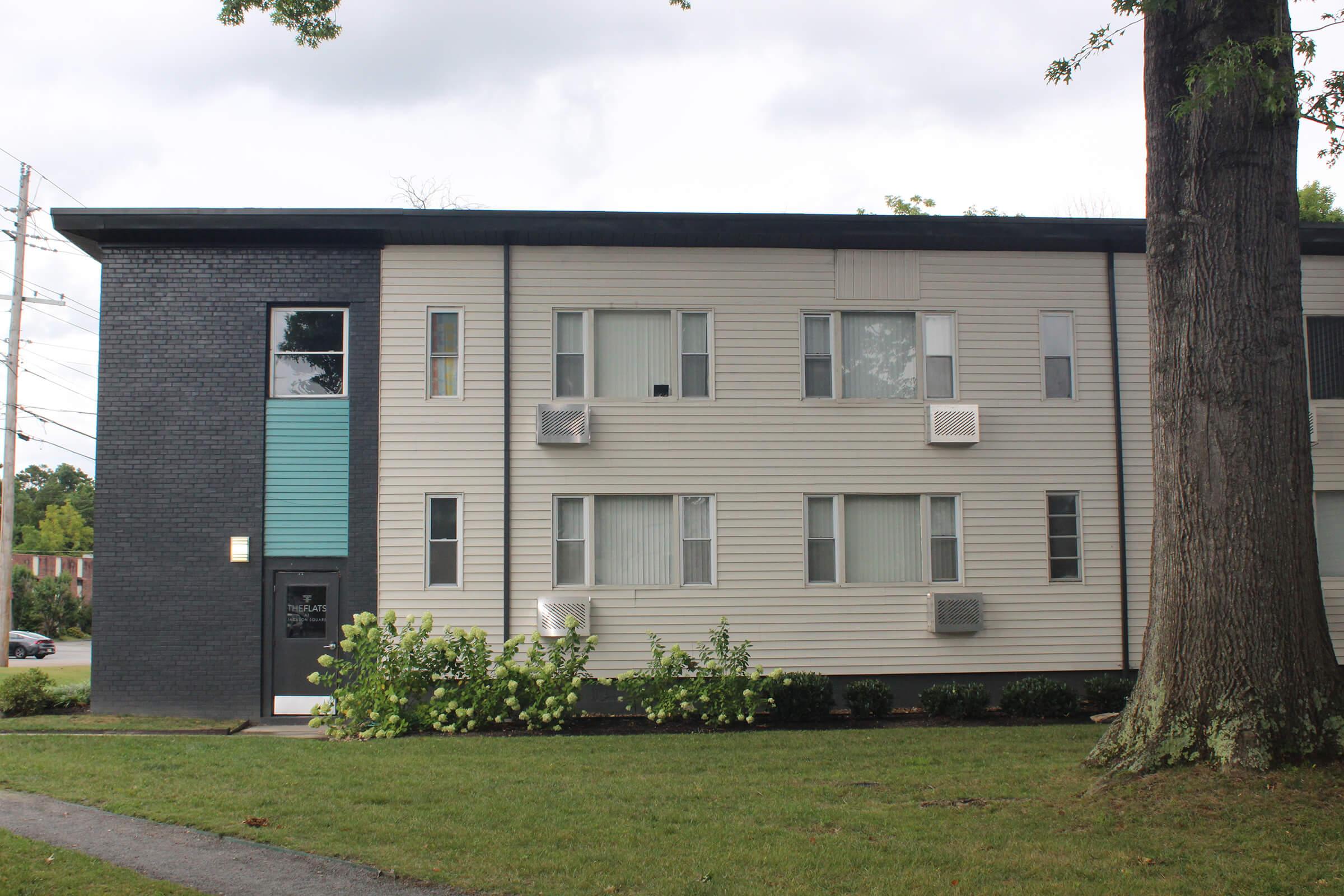 A multi-story apartment building featuring a mix of light-colored siding and dark brick. The facade includes several windows, some with air conditioning units, and an entrance door on the left side. A grassy area with small shrubs is visible in front, along with a tree on the right.