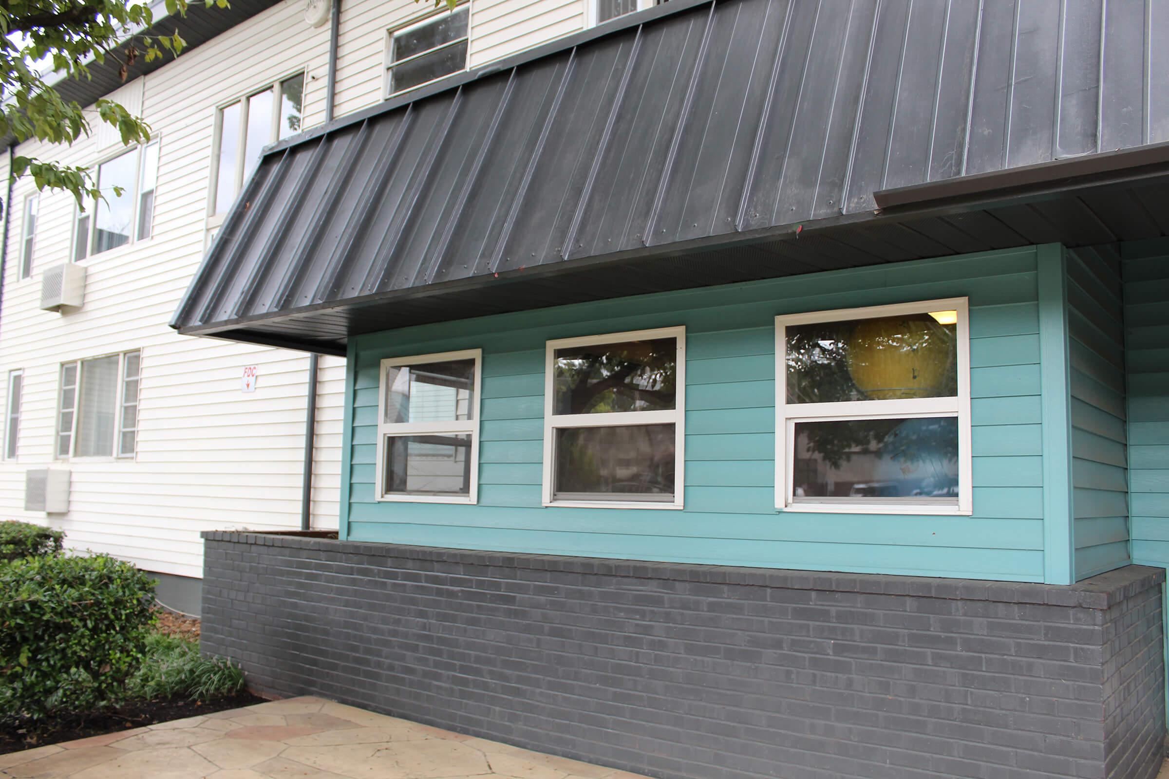 A building with a two-tone exterior featuring a turquoise wall section and large windows below a black metal awning. The structure has both light and dark elements, with a neatly paved area in front and landscaped greenery nearby.