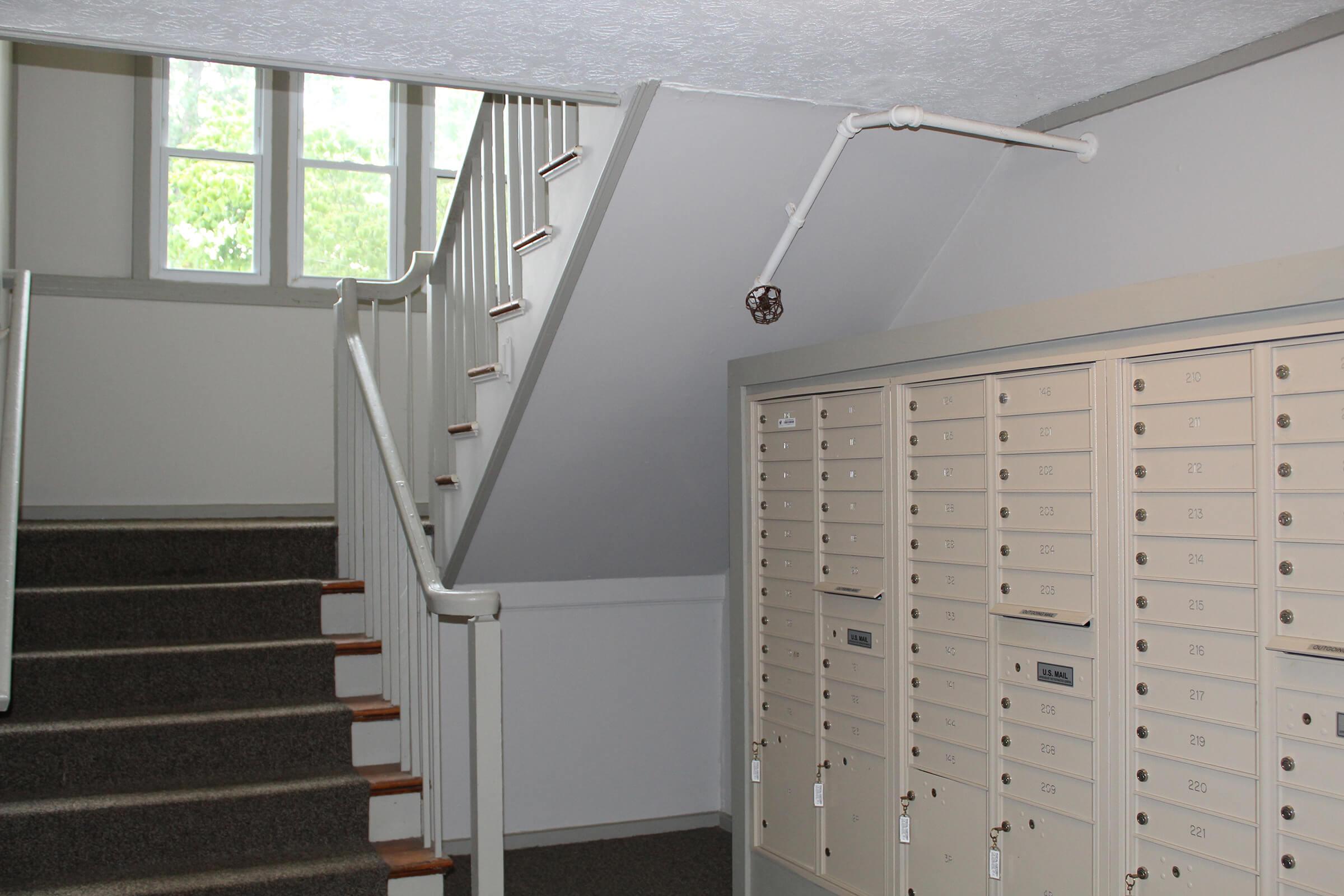 Indoor scene featuring a staircase leading to an upper level. On the right, there is a wall-mounted mailbox unit with multiple compartments, while the left side shows the carpeted stairs and a portion of a handrail. Natural light filters through windows at the top of the staircase, illuminating the area.