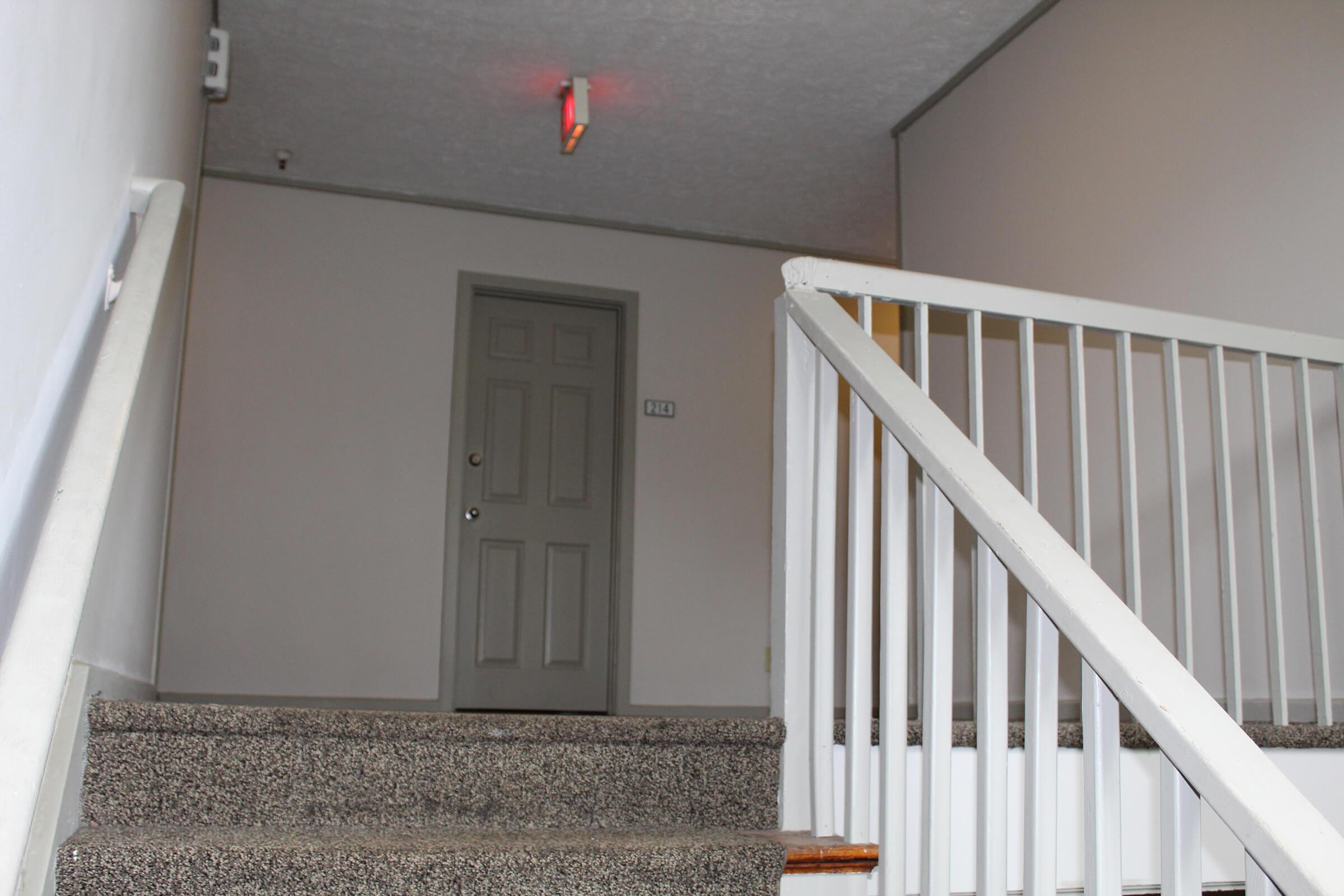A stairway leading to a closed door (numbered 212) at the top of the stairs. The walls are painted a light color, and a red light is visible in the ceiling, indicating the hallway's light fixture. The railing is white, and the stairs are carpeted with a speckled pattern.