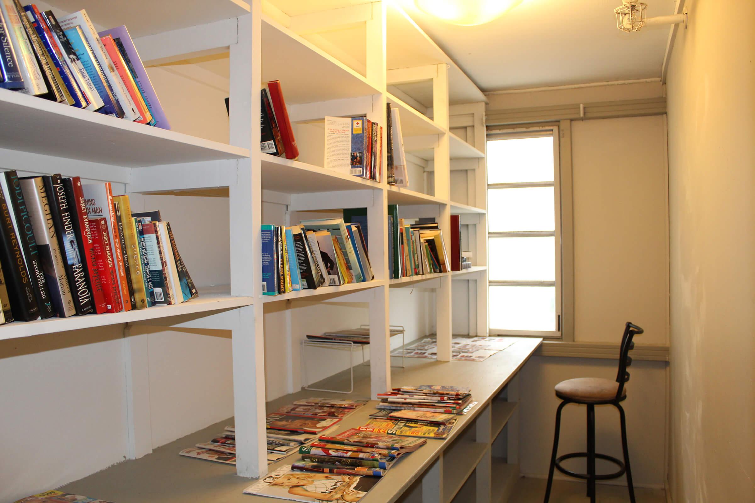 A cozy reading nook with white shelves filled with various books. A desk is positioned against the wall, adorned with magazines. A simple chair is placed at the desk, and a window allows natural light to brighten the space. The overall atmosphere is inviting and conducive to reading or studying.