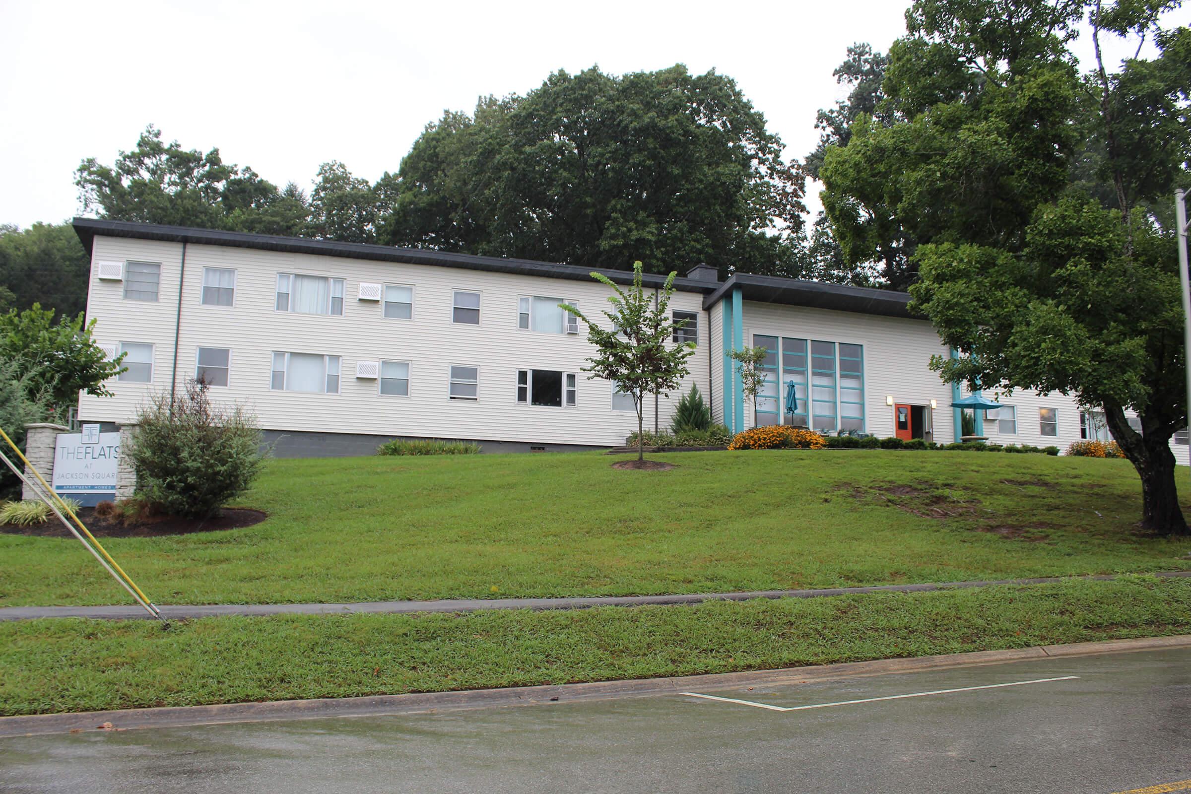 A two-story residential building surrounded by greenery, featuring a large front window and multiple windows along the sides. The area is well-maintained with shrubs and flowers, and there is a sign visible that reads "The Flats." The sky is overcast, and the ground is wet, suggesting recent rain.