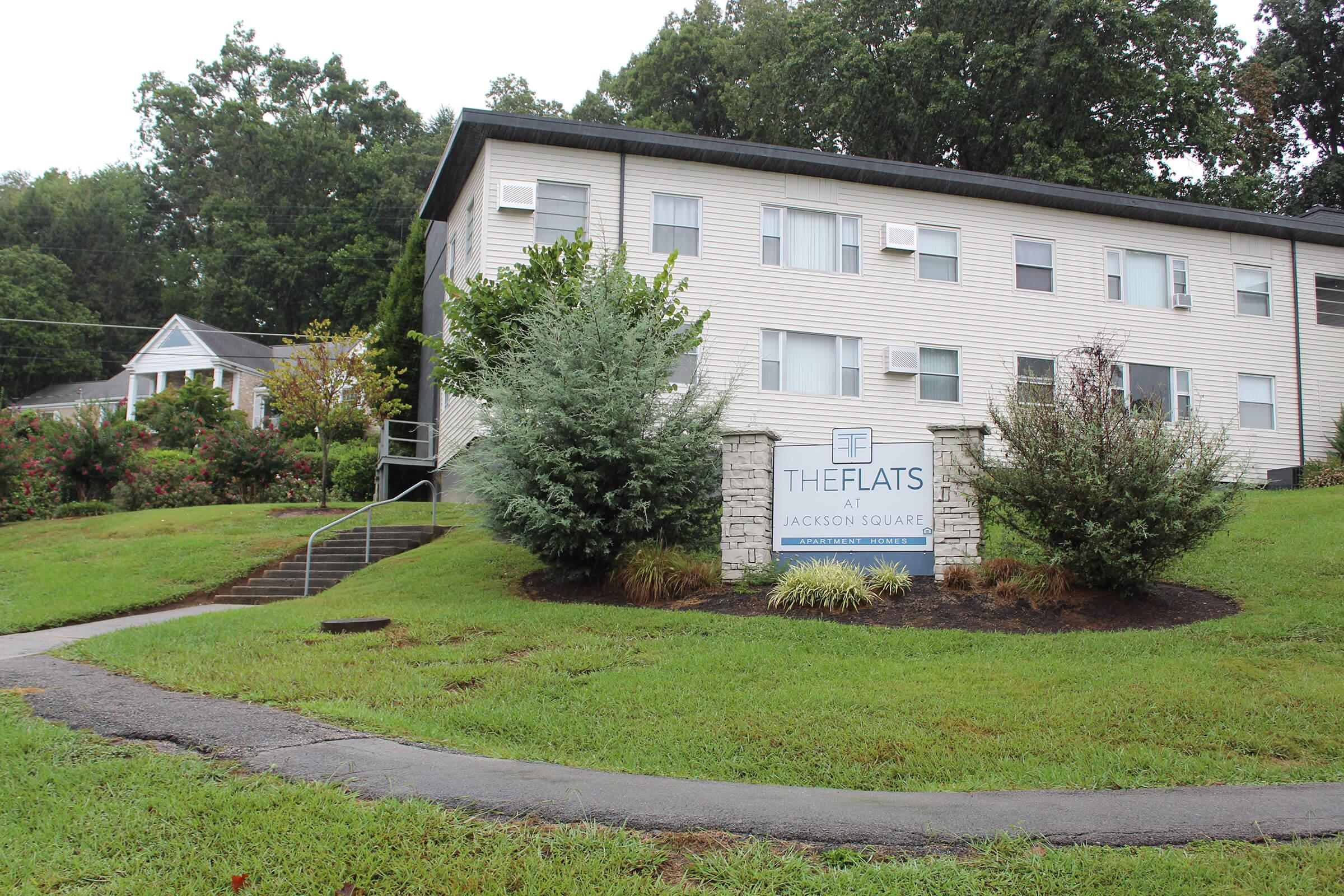 The image shows a residential apartment building named "The Flats at Jackson Square," set in a green landscape with manicured bushes. A sign is prominently displayed in front of the building. There are stairs leading up to the entrance, and the scene is overcast, indicating cloudy weather.
