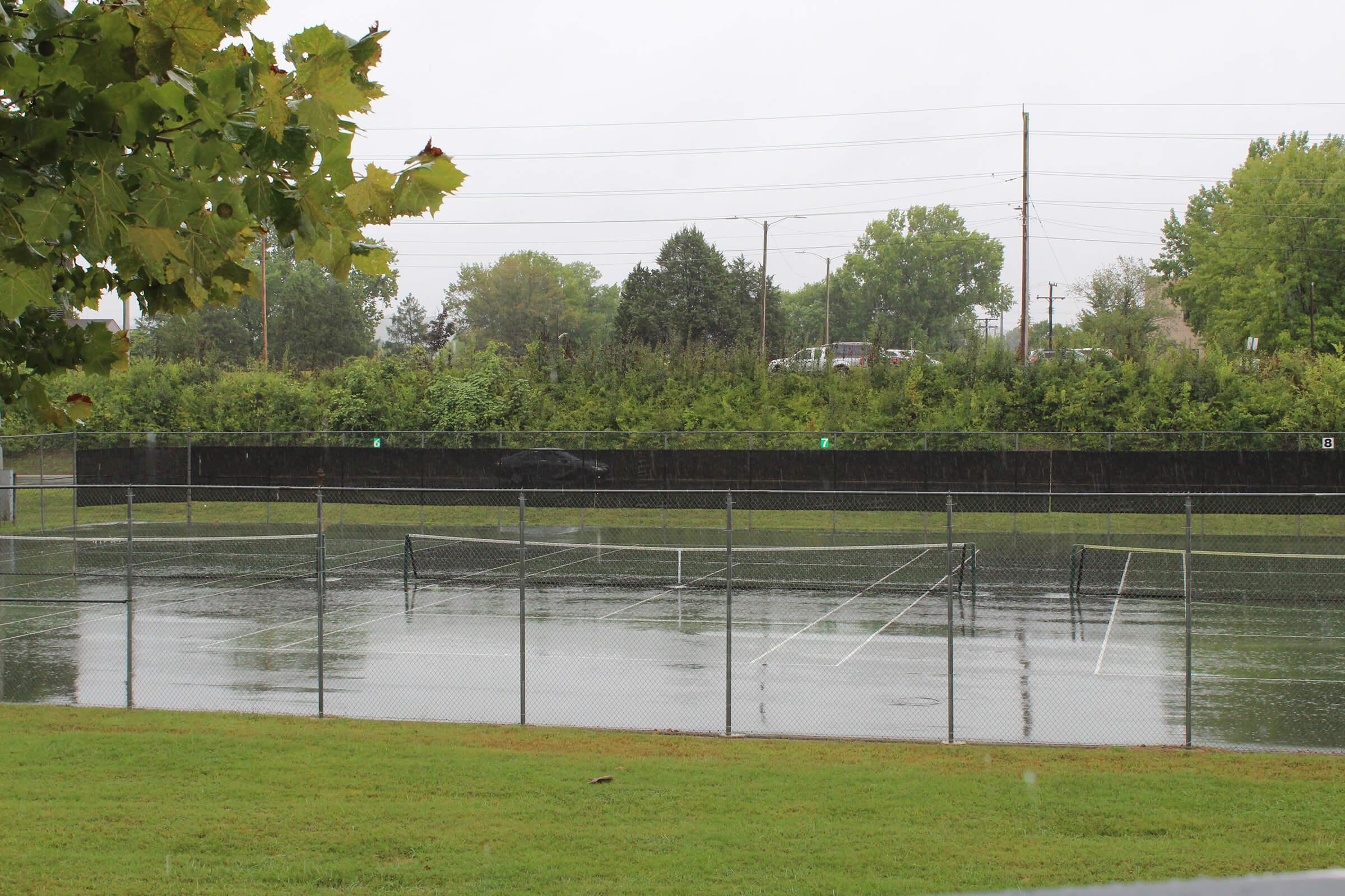 A rain-soaked tennis court surrounded by green grass and trees, with water pooling on the surface. The sky is overcast, and the scene conveys a gloomy, rainy day.