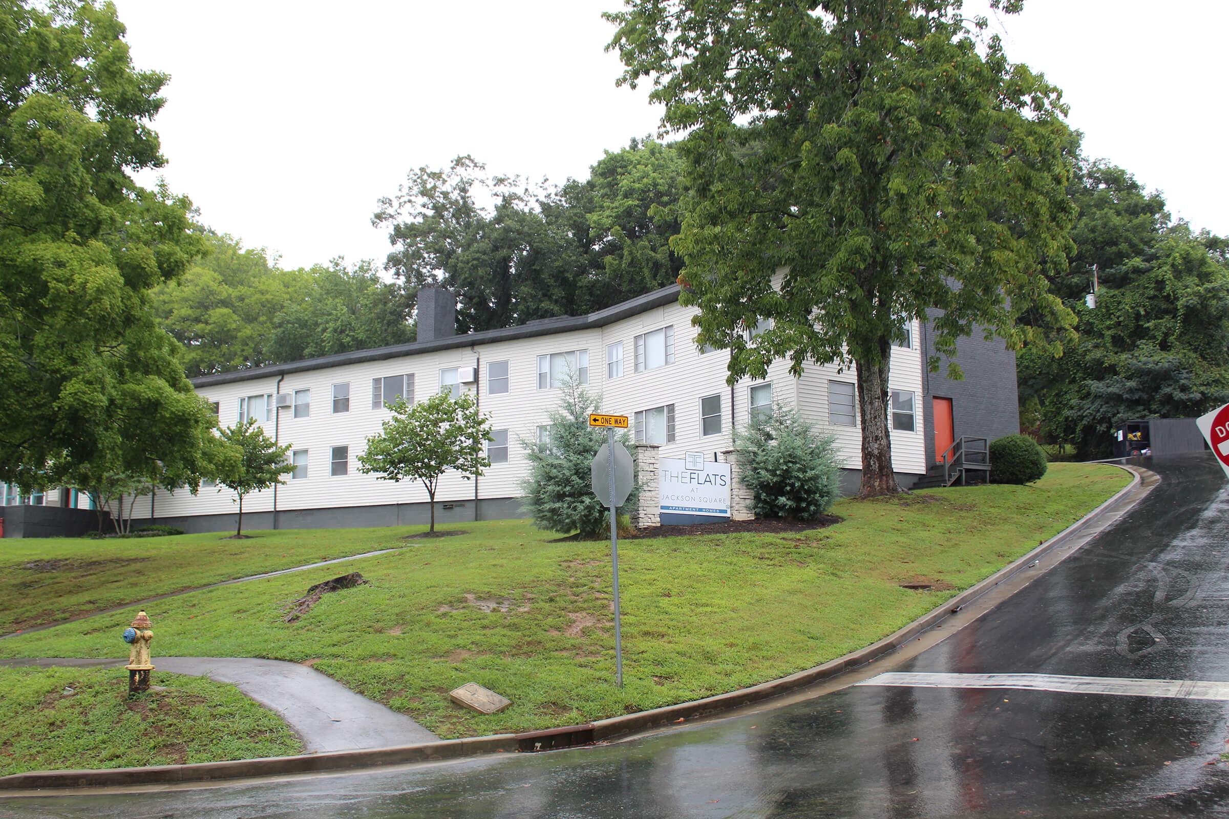 A low-rise apartment building with light-colored siding, surrounded by greenery and trees. There are two small trees planted in front of the building, and a fire hydrant is visible on the sidewalk. The street is wet, suggesting recent rain, and a stop sign is present at the corner of the road.