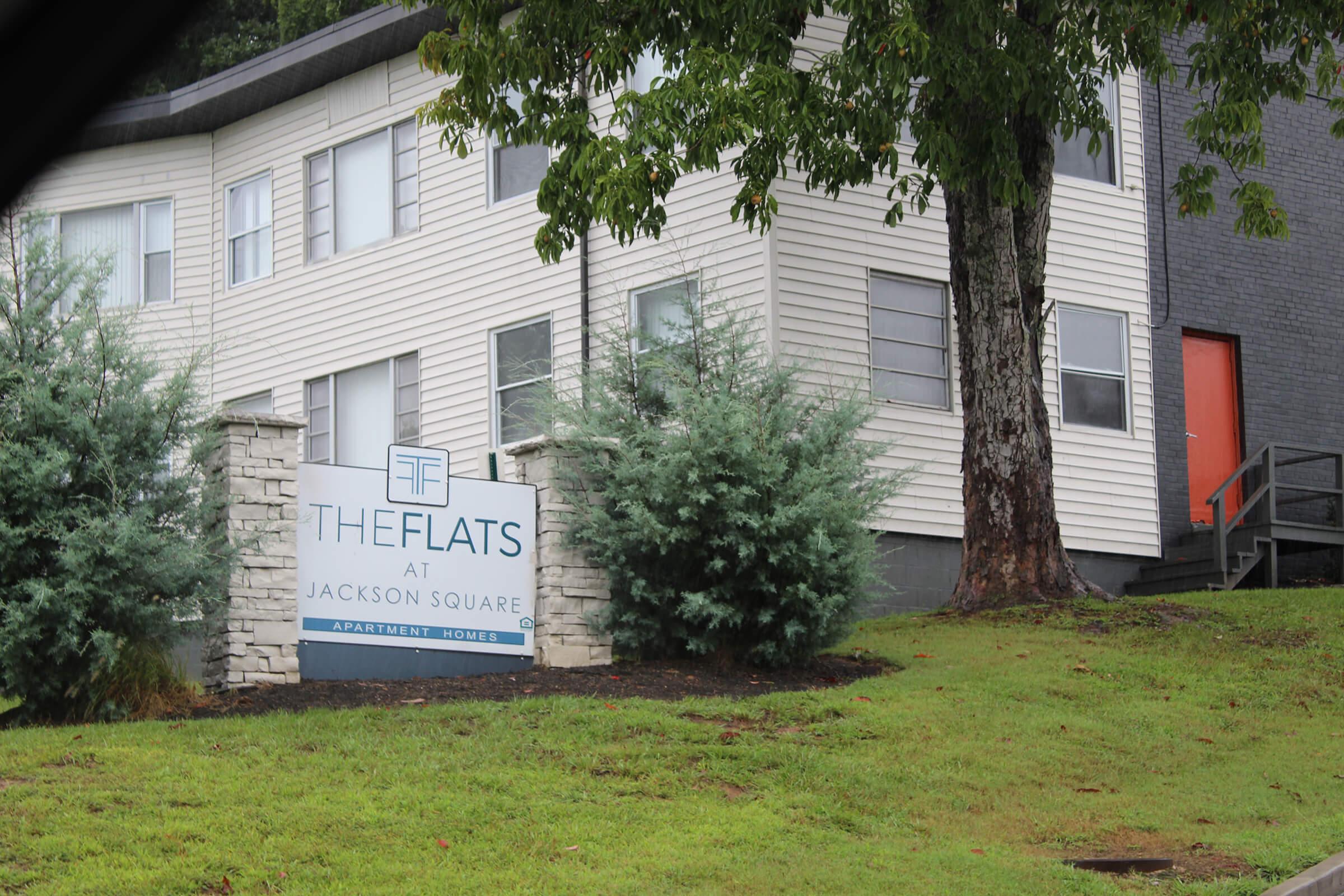 Apartment complex sign for "The Flats at Jackson Square," featuring a modern design with green shrubbery in front. The building has a two-tone exterior with a distinctive orange door visible. The setting includes a grassy area and landscaping elements.