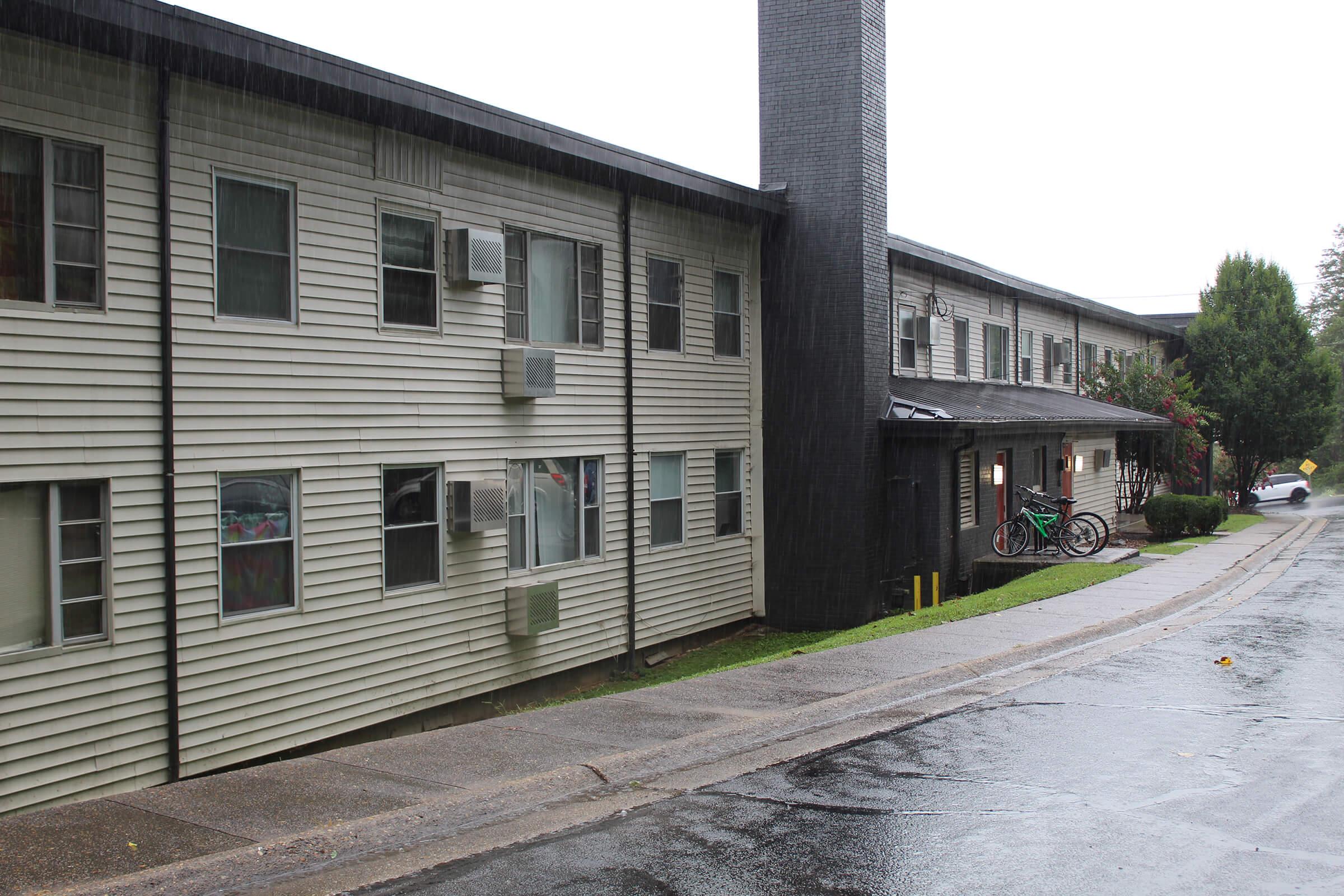 A view of a residential building on a rainy day, featuring a two-story structure with several air conditioning units on the exterior. A narrow road runs alongside the building, and bicycles are visible near the entrance. Wet pavement reflects the overcast sky, adding to the somber atmosphere.