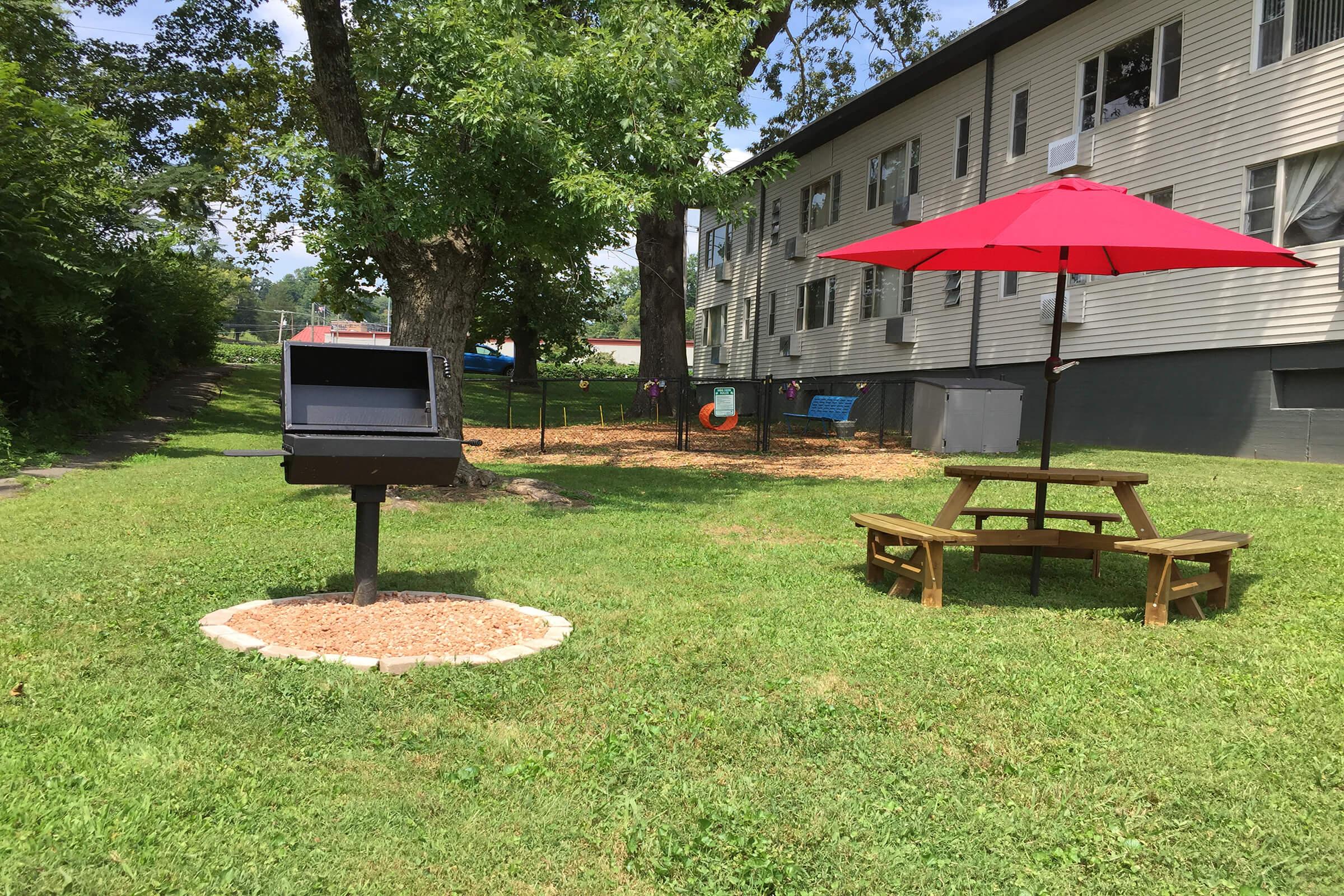 A grassy area with a black grill station on a stone base, a wooden picnic table, and a bright red umbrella. In the background, there is a playground with swings and slides, along with a multi-story building. Lush trees provide shade in the sunny outdoor space.