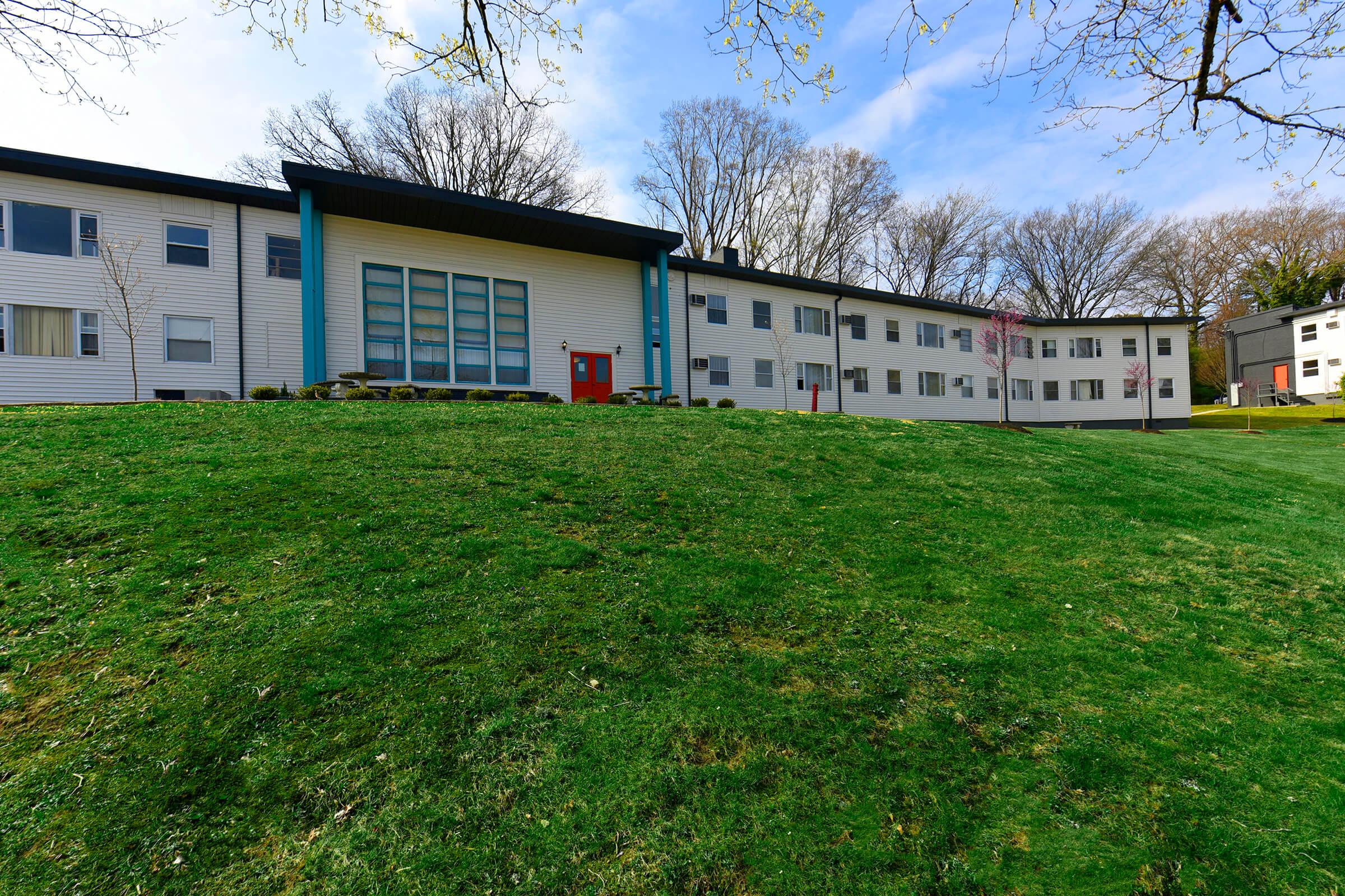 A modern, multi-story building featuring a large front entrance with large windows and a bright red door, set on a green lawn. The building has a white exterior with some trees in the background and a clear blue sky overhead.