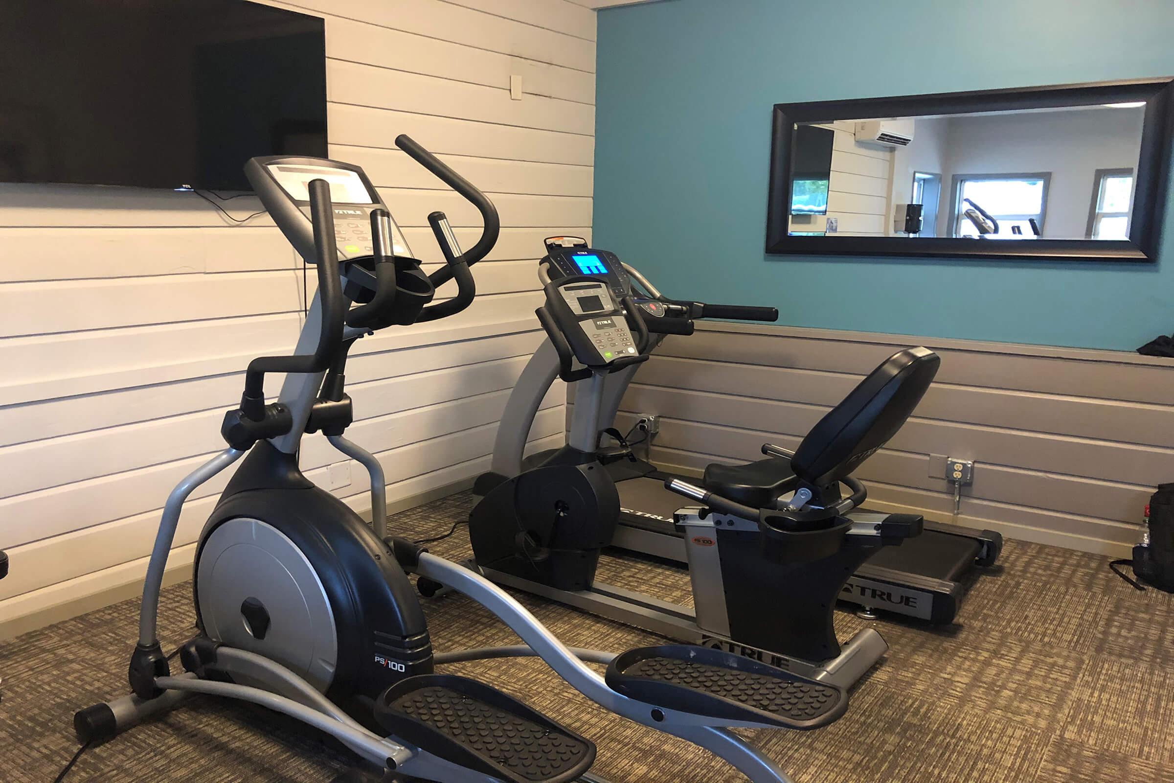 A small, well-lit fitness room featuring an elliptical machine and a recumbent bike. A television is mounted on the wall, and there's a mirror reflecting the equipment. The floor is carpeted, and the walls are painted blue and white, creating a fresh atmosphere for exercise.