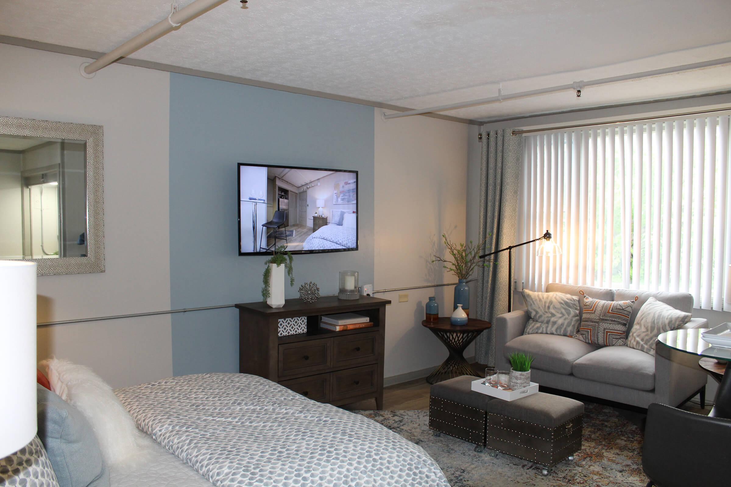 A cozy living space featuring a comfortable gray sofa with decorative pillows, a coffee table with a plant, and a dresser. A flat-screen TV is mounted on the wall displaying a bedroom image. There are curtains letting in natural light, with a mirror reflecting part of the room. Neutral colors create a calm atmosphere.
