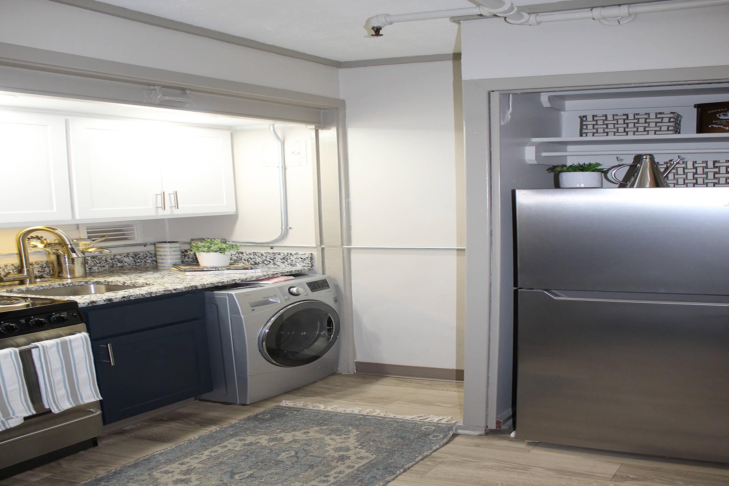 A modern kitchen area featuring a stainless steel refrigerator and a washing machine. The kitchen has white and dark blue cabinetry, a granite countertop, and a decorative rug on the floor. There are small plants and storage boxes on the shelves, creating a tidy and stylish space.