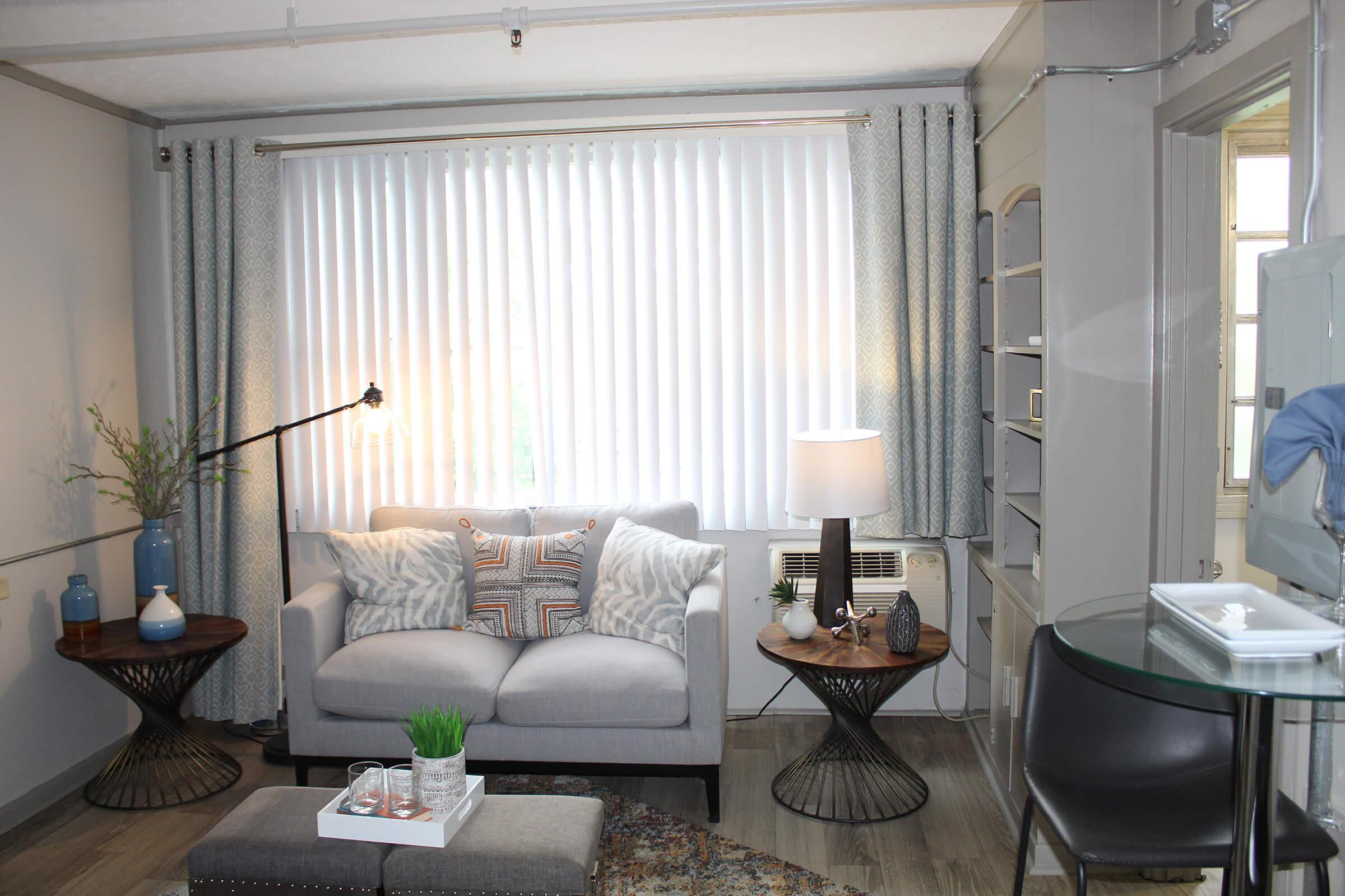 A cozy living room featuring a light gray sofa with decorative pillows, two round side tables with vases and plants, a standing lamp, and white vertical blinds covering a window. An air conditioning unit is visible, along with a small rug on the floor and a glass-top table in the corner.