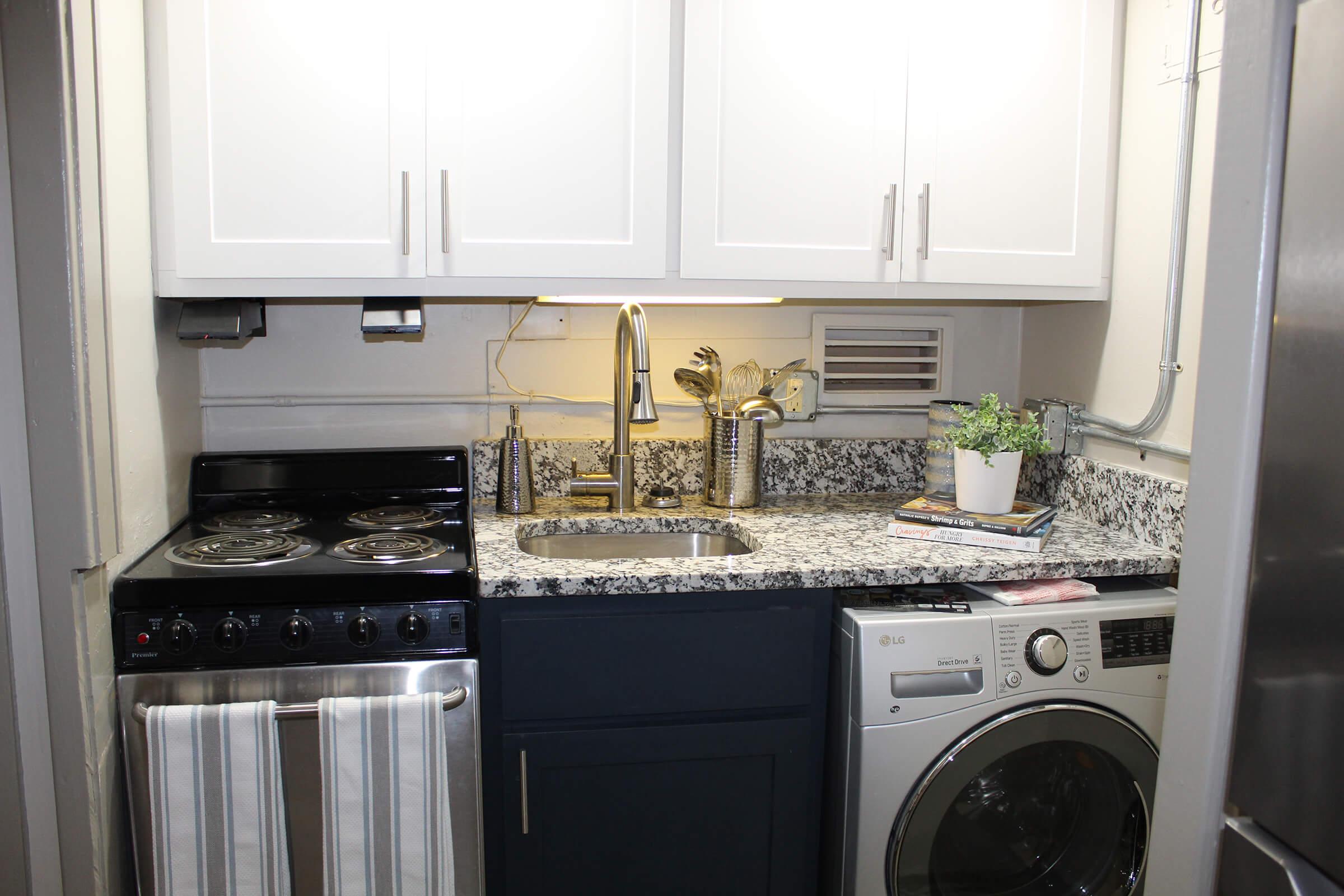A compact laundry area featuring a stacked washer and dryer, a black stove, and a double sink with a modern faucet. The counter is made of speckled granite, with white cabinets above and a small plant and magazines on the countertop, providing a tidy and functional space.
